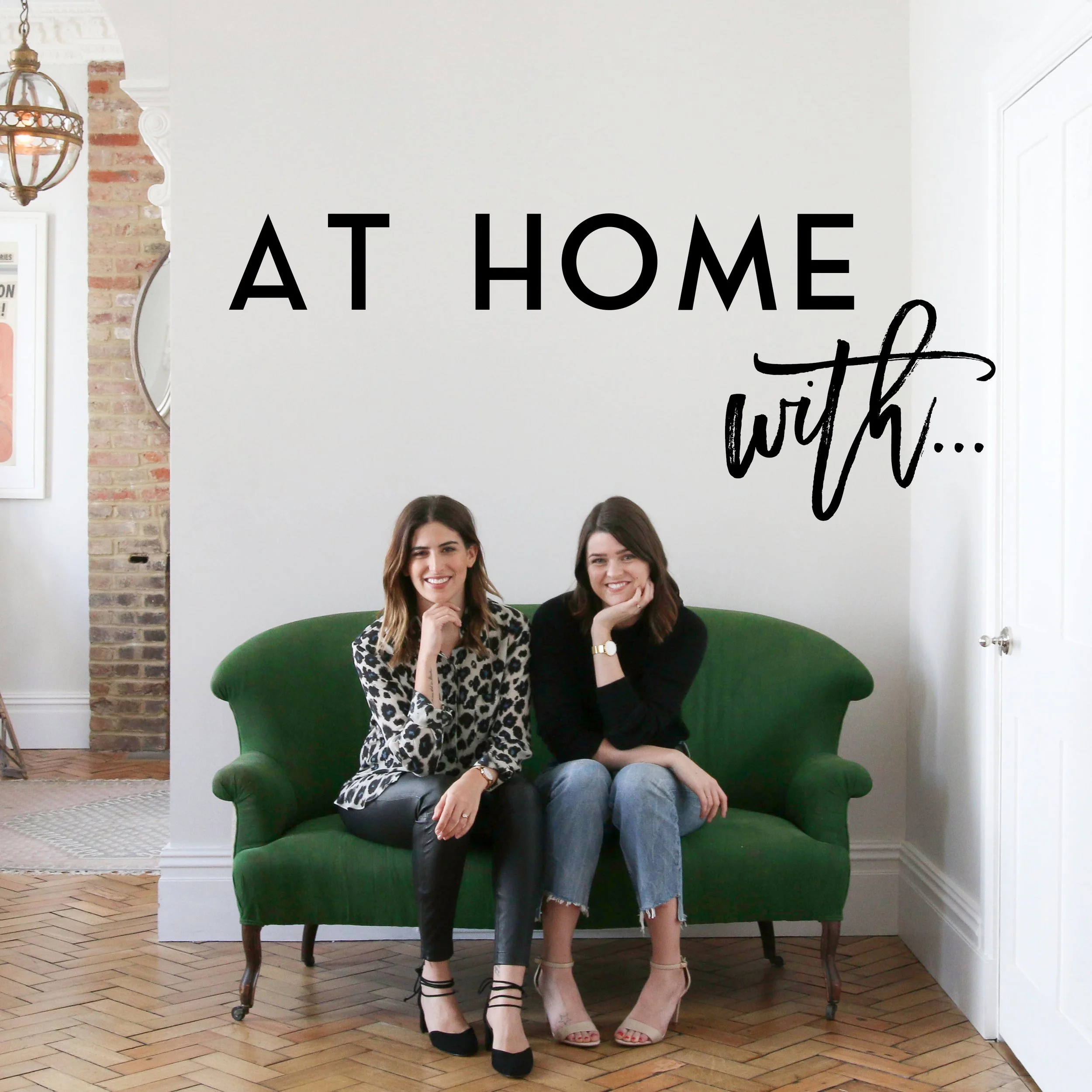 Two women sitting on a green sofa in a bright, modern room with hardwood floors, smiling at the camera. There is a wall decal that reads 'At Home with...' in black text. One woman is wearing a leopard print top and black pants, while the other is wearing a black top and ripped jeans.