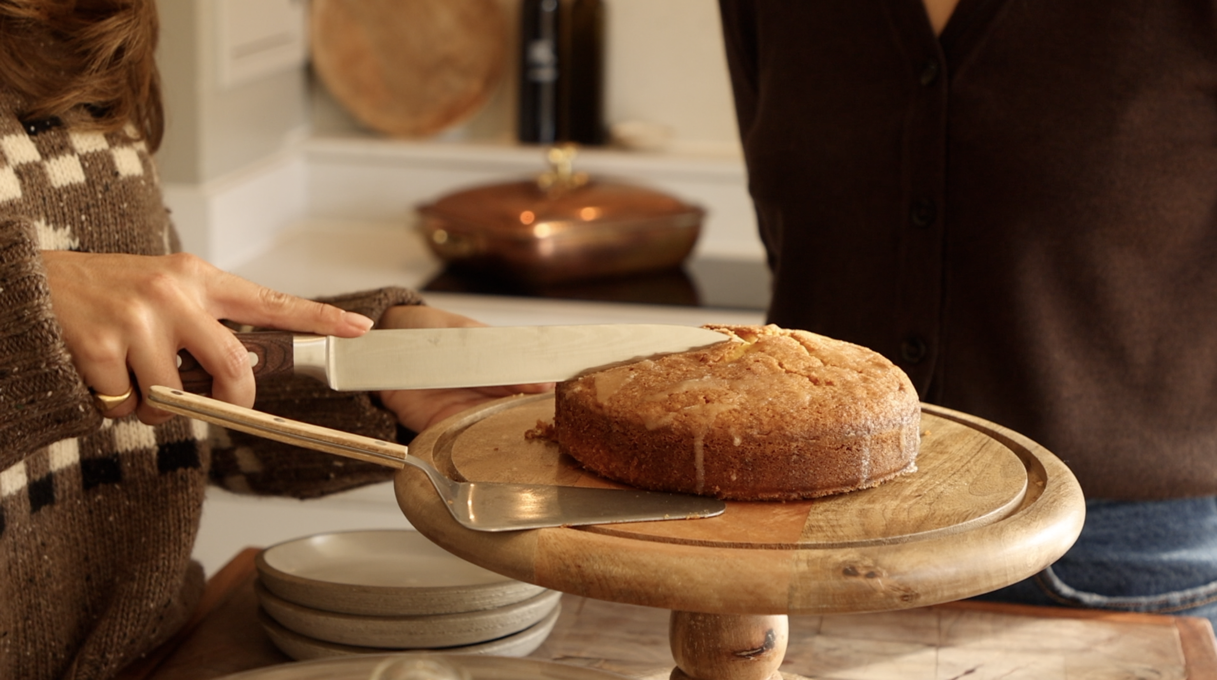 Person slicing a freshly baked cake on a wooden cake stand in a kitchen.