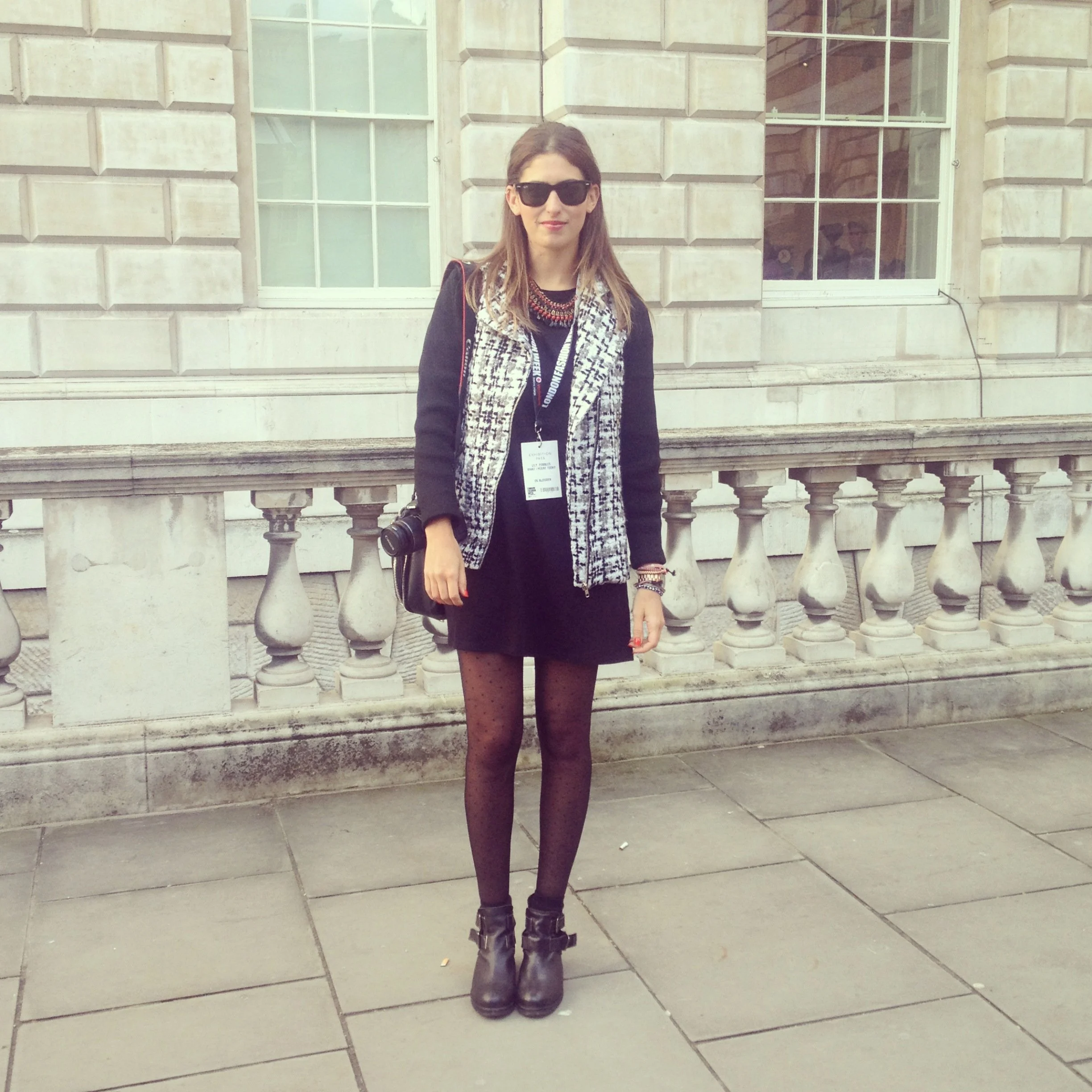 A woman standing on a sidewalk in front of a historic building with a stone balustrade, wearing black sunglasses, a black dress, patterned tights, ankle boots, and a patterned coat, holding a camera and wearing a badge around her neck.