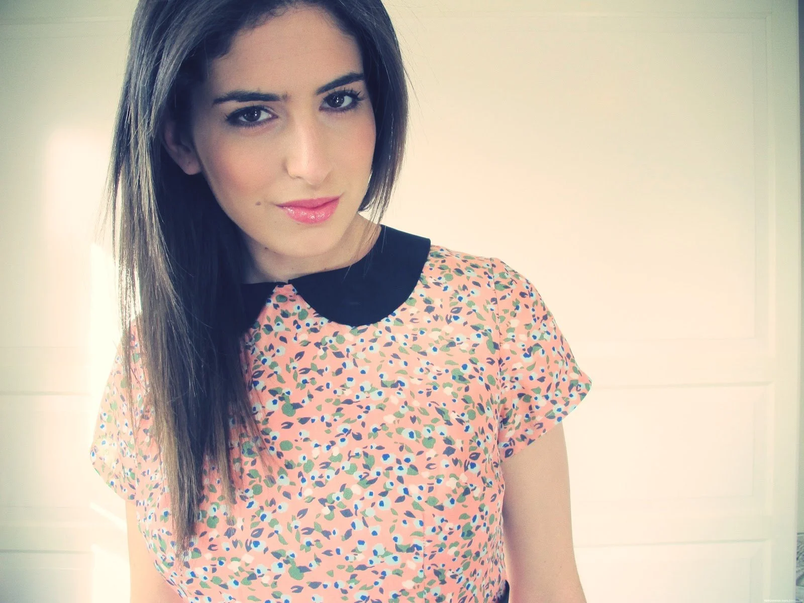 A young woman with shoulder-length brown hair and light makeup, wearing a short-sleeved printed pink shirt with a black collar, standing indoors with a plain light-colored wall in the background.
