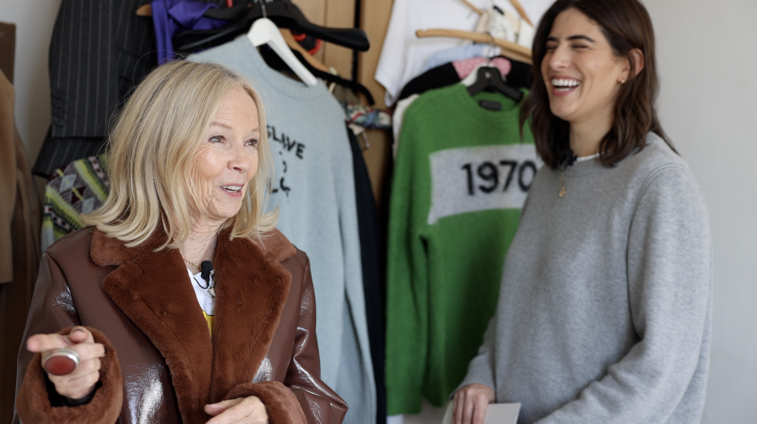 Two women chatting and smiling in a clothing store surrounded by sweaters and jackets hanging on the wall.