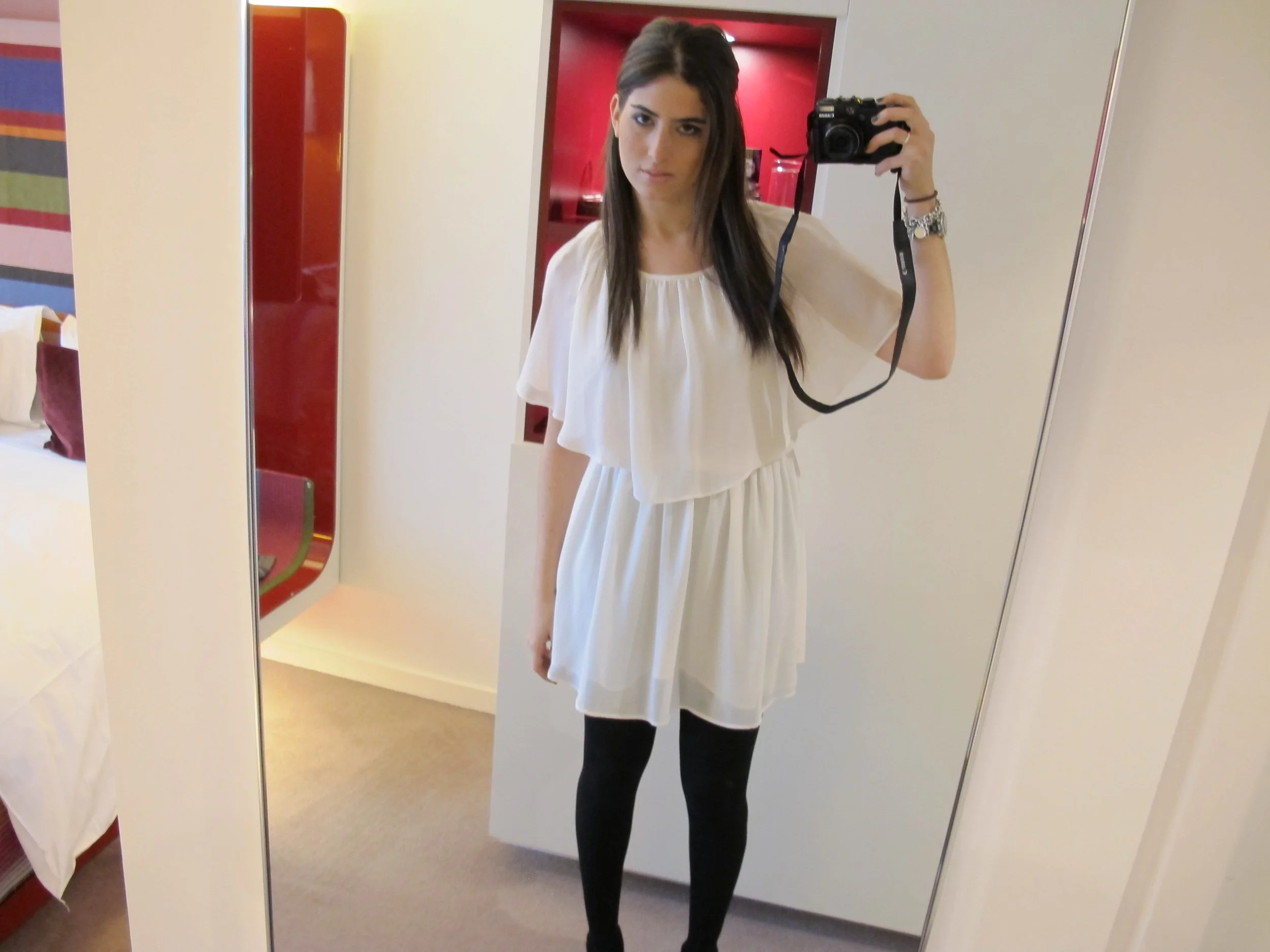 A young woman taking a selfie in a hotel room mirror. She has long dark hair, is wearing a white dress and black tights, and has a camera strap around her wrist.