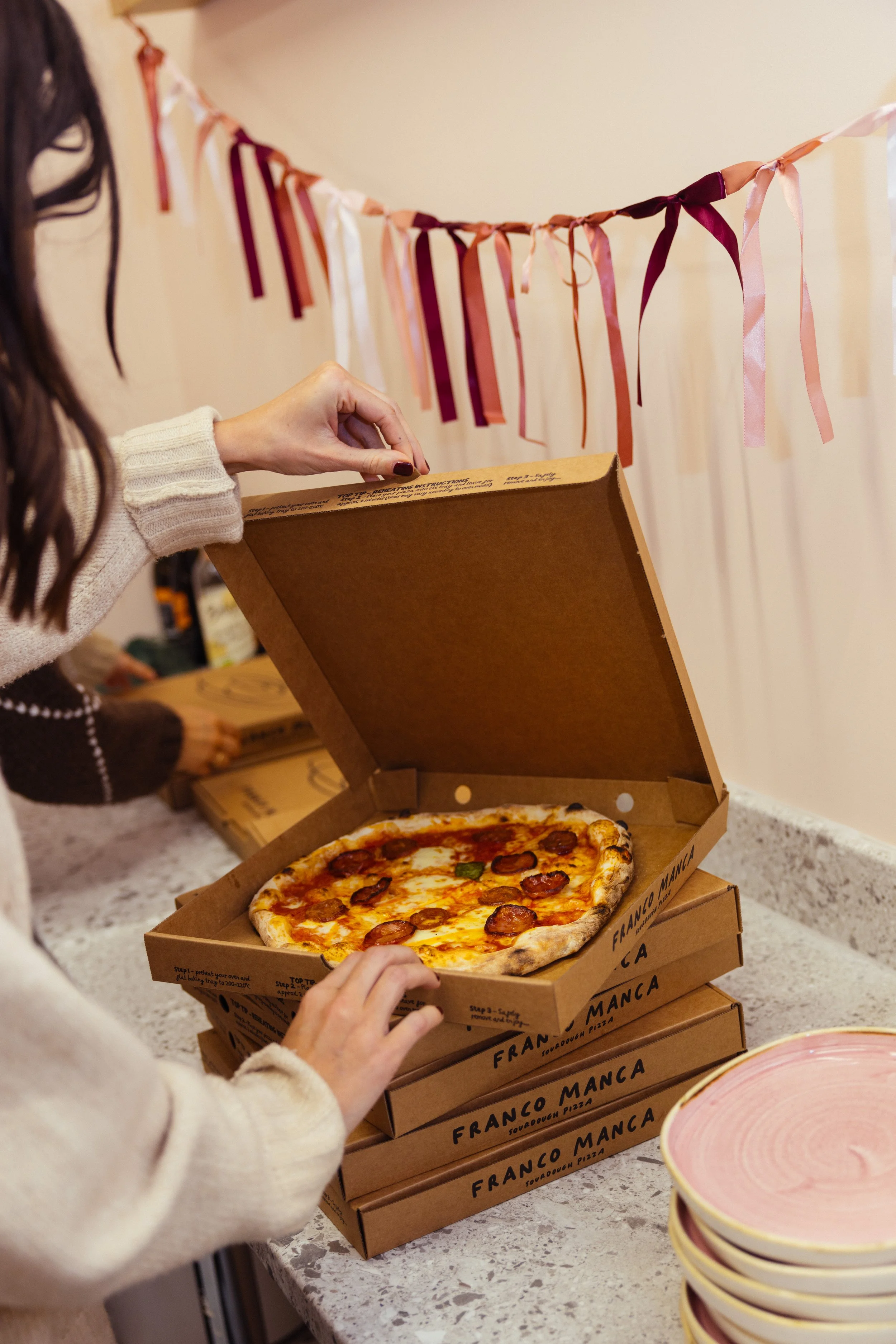 Person opening a pizza box with a pepperoni and green pepper pizza inside, with additional pizza boxes stacked nearby, in front of a decorative pink and white ribbon on the wall.