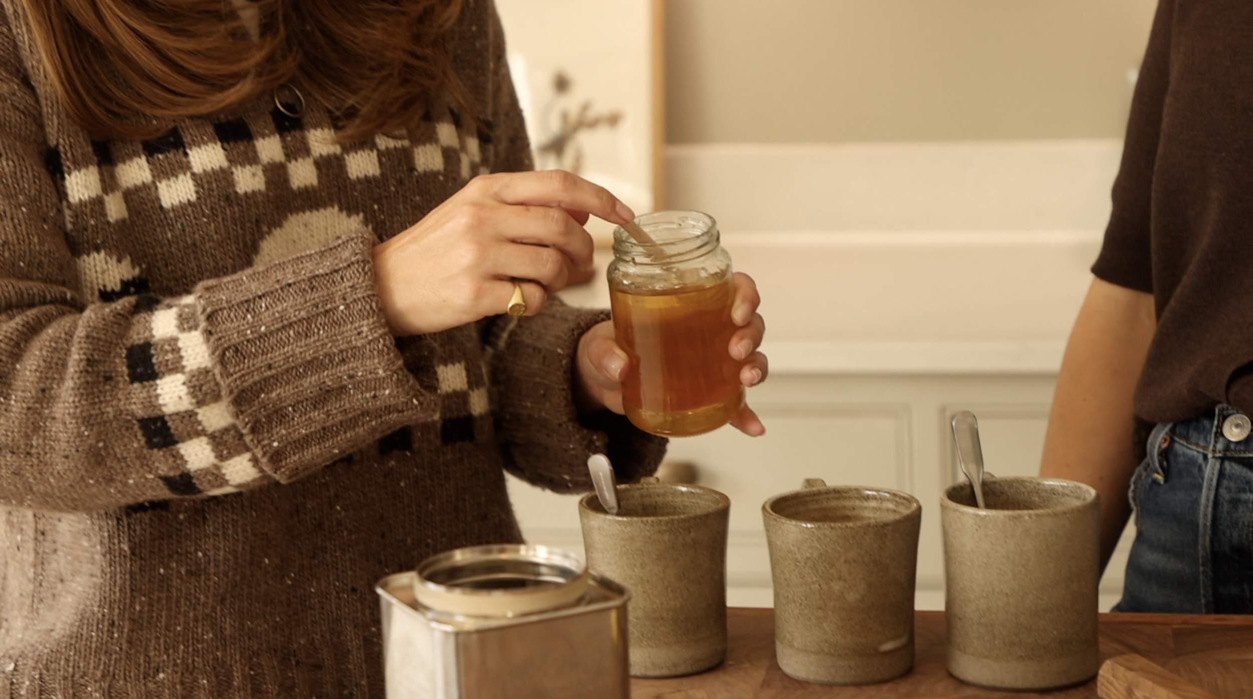 A woman in a brown patterned sweater is holding a jar of honey with a honey dipper, pouring honey into three ceramic cups with spoons in them, on a wooden table.
