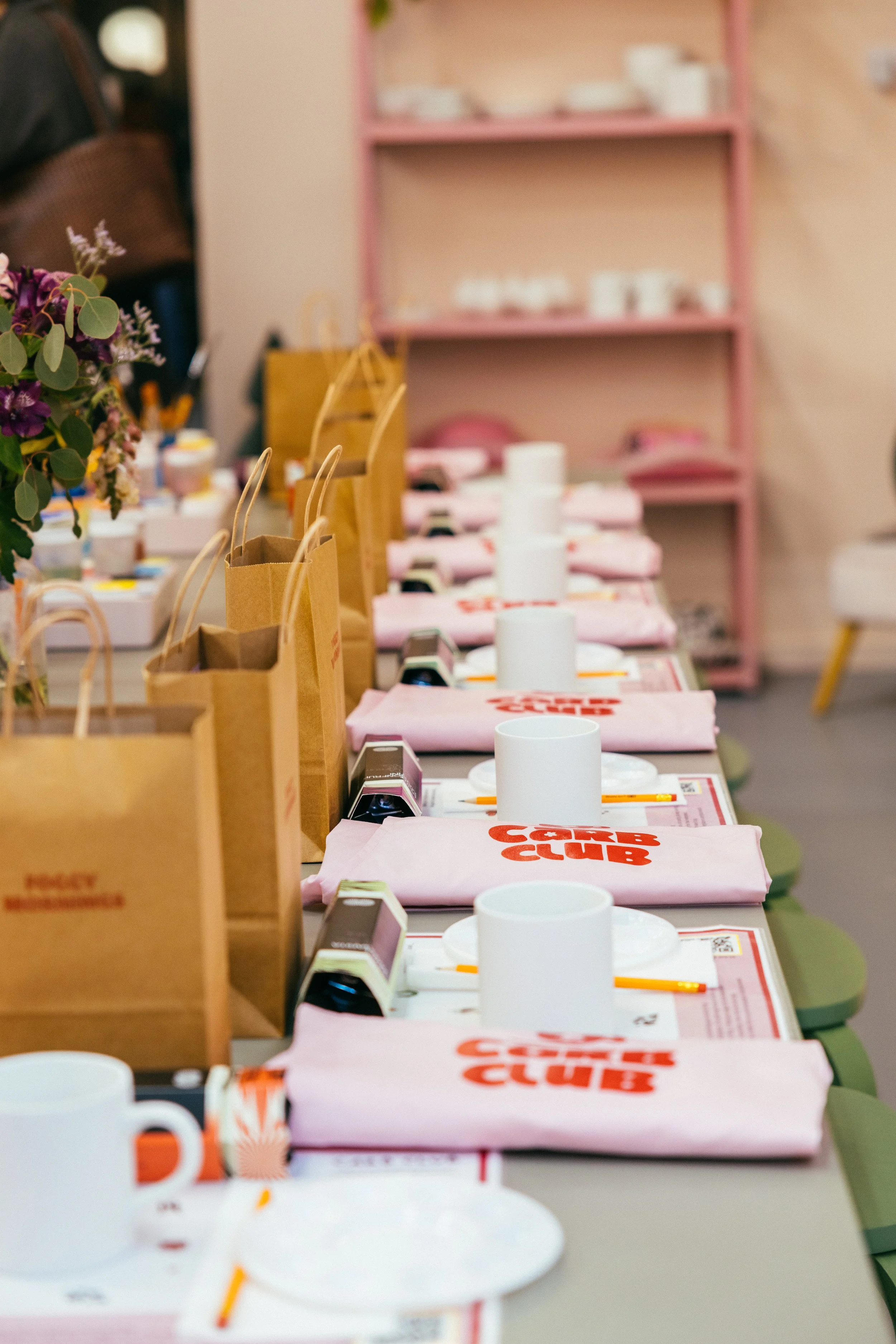 A row of tables set up for a corporate event with pink tote bags, white cups, paper napkins, and organization materials, with a pink shelf in the background.