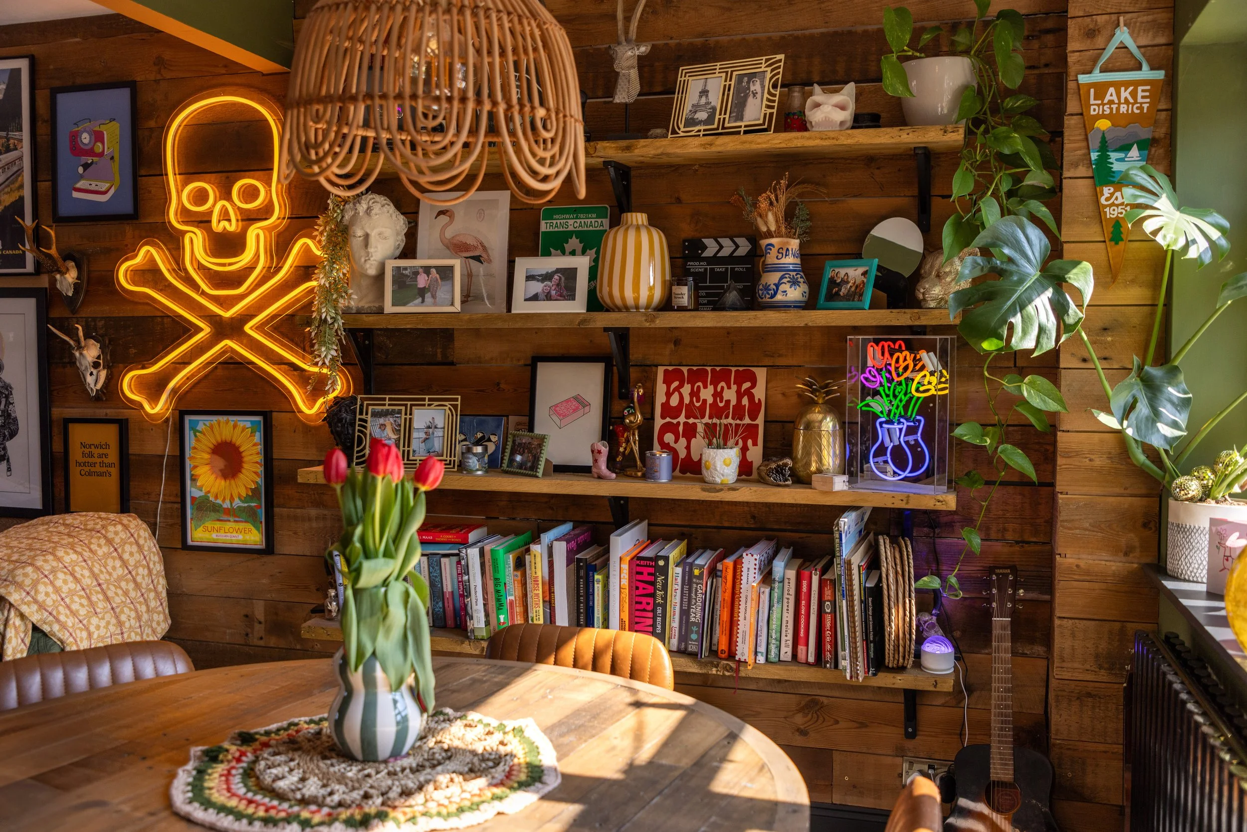 A cozy living room corner with wood-paneled walls decorated with various framed pictures, a neon skull and crossbones sign, and a large neon vase with flowers. A wooden bookshelf holds books and decorative items, and a guitar leans against the wall. A round table in the foreground has a colorful, crocheted tablecloth and a vase of red tulips. The space is filled with plants and warm lighting.
