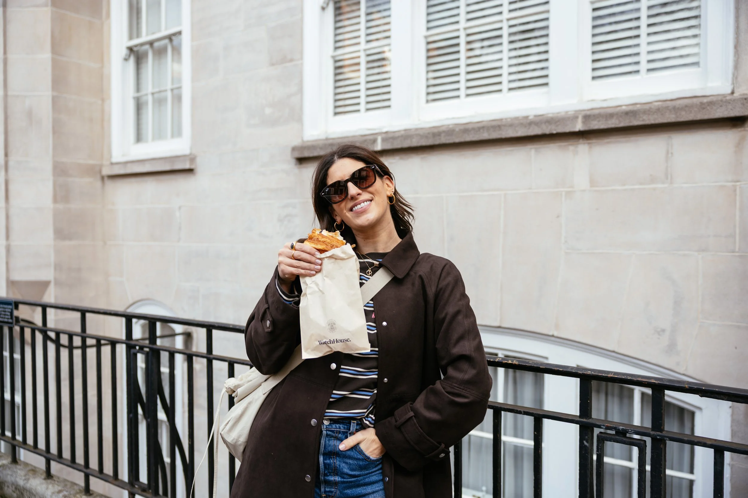 A woman wearing sunglasses and a dark coat holding a paper bag with a pastry outside in an urban area.