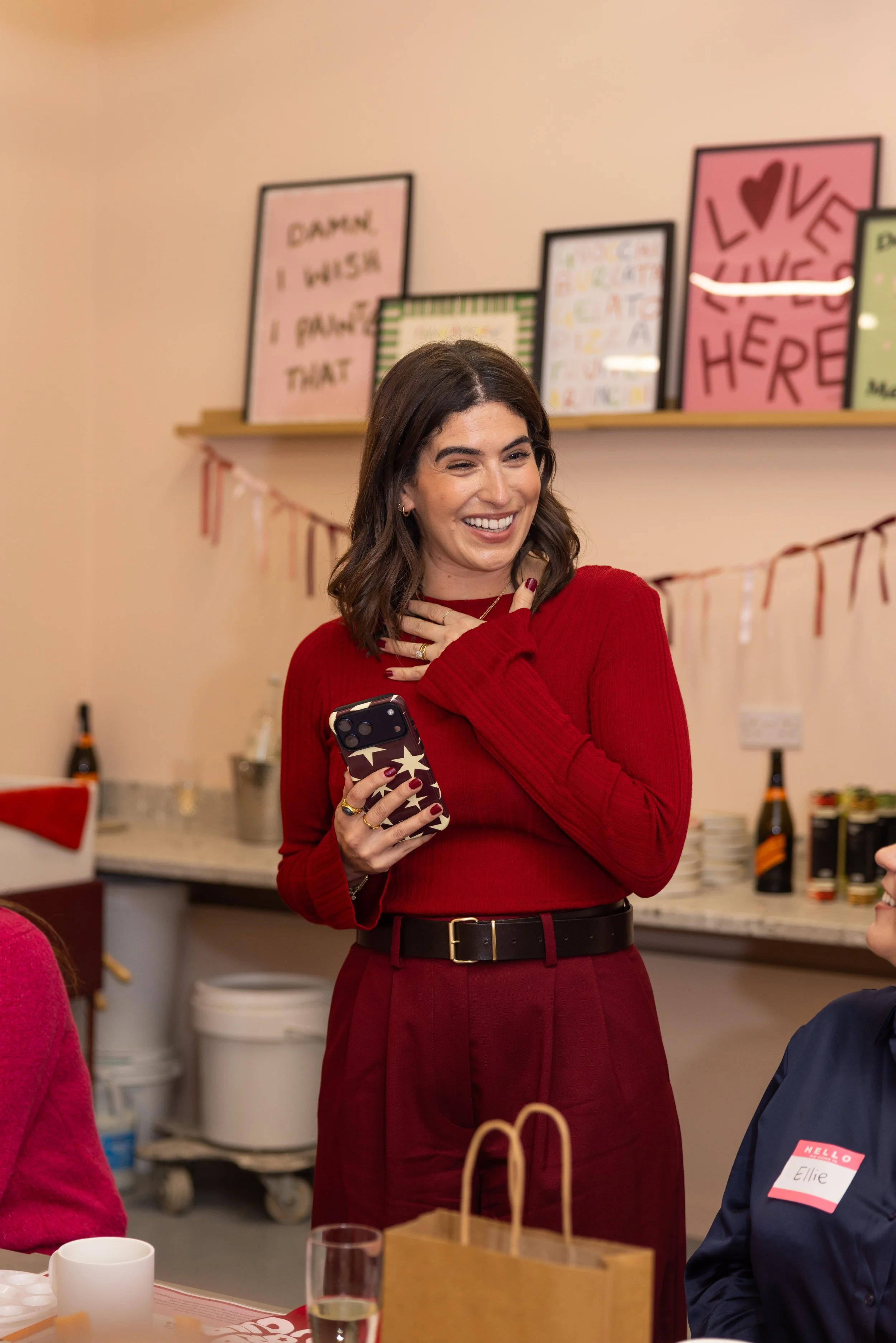 A woman in a red sweater and dark red pants holding a phone, smiling and touching her chest, at a party or gathering with decorations and drinks.