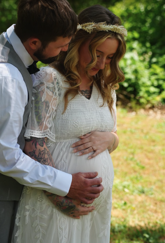 A pregnant woman in a white dress with lace details, wearing a floral headband, standing outdoors with a man embracing her from behind, both with their hands on her belly, in a lush green setting.