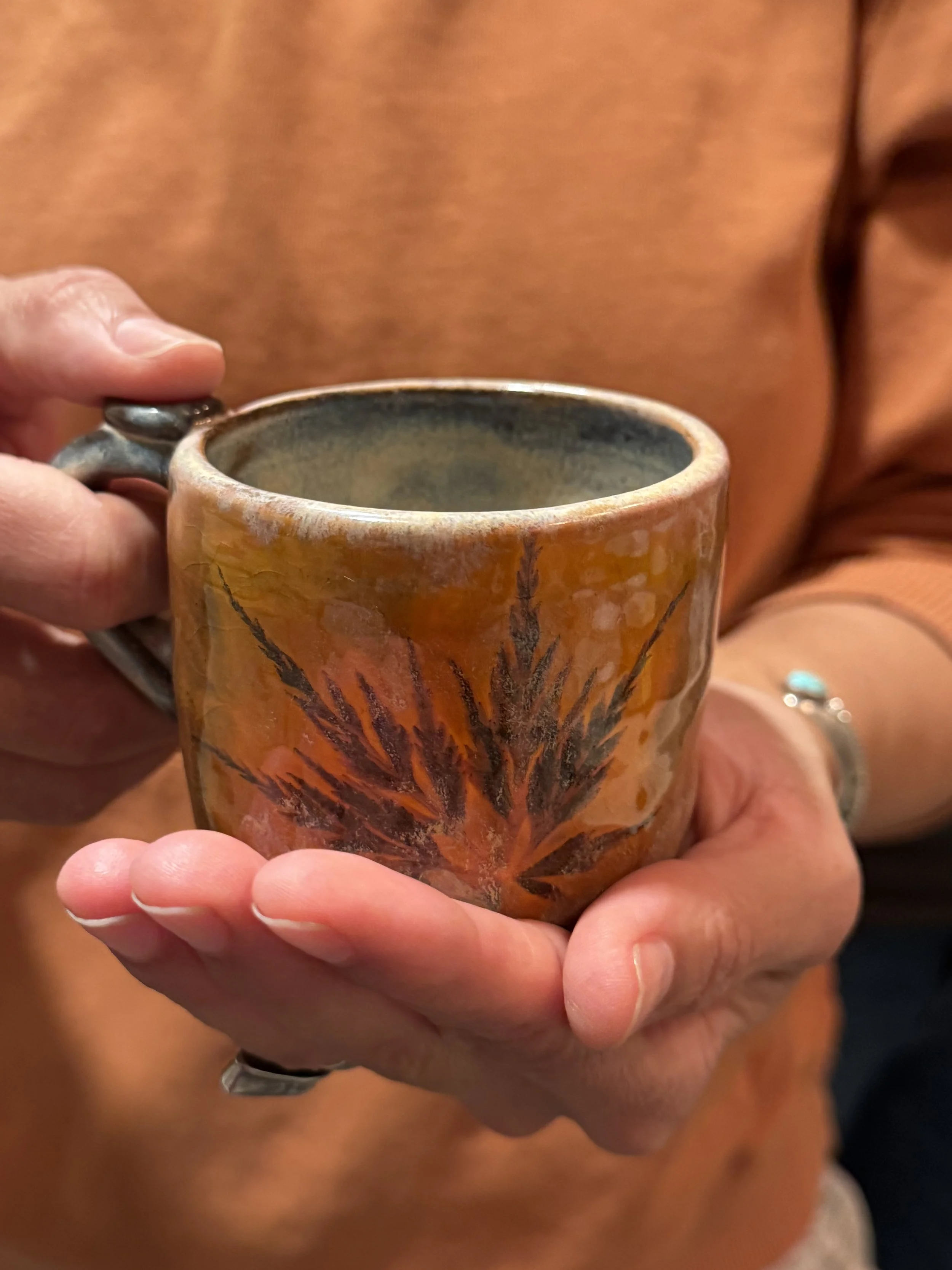 Close-up of a person holding a ceramic mug with a leaf design, wearing an orange shirt and a bracelet with turquoise stones.