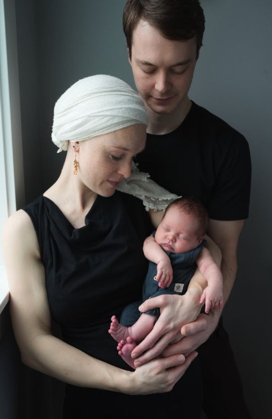 A woman with a white headscarf holding a sleeping baby, with a man standing behind her gently supporting them.