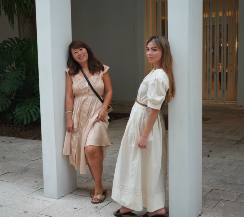 Two women in light-colored dresses standing at a white architectural opening, with one smiling and the other looking serious, in an outdoor setting with greenery and a modern building in the background.