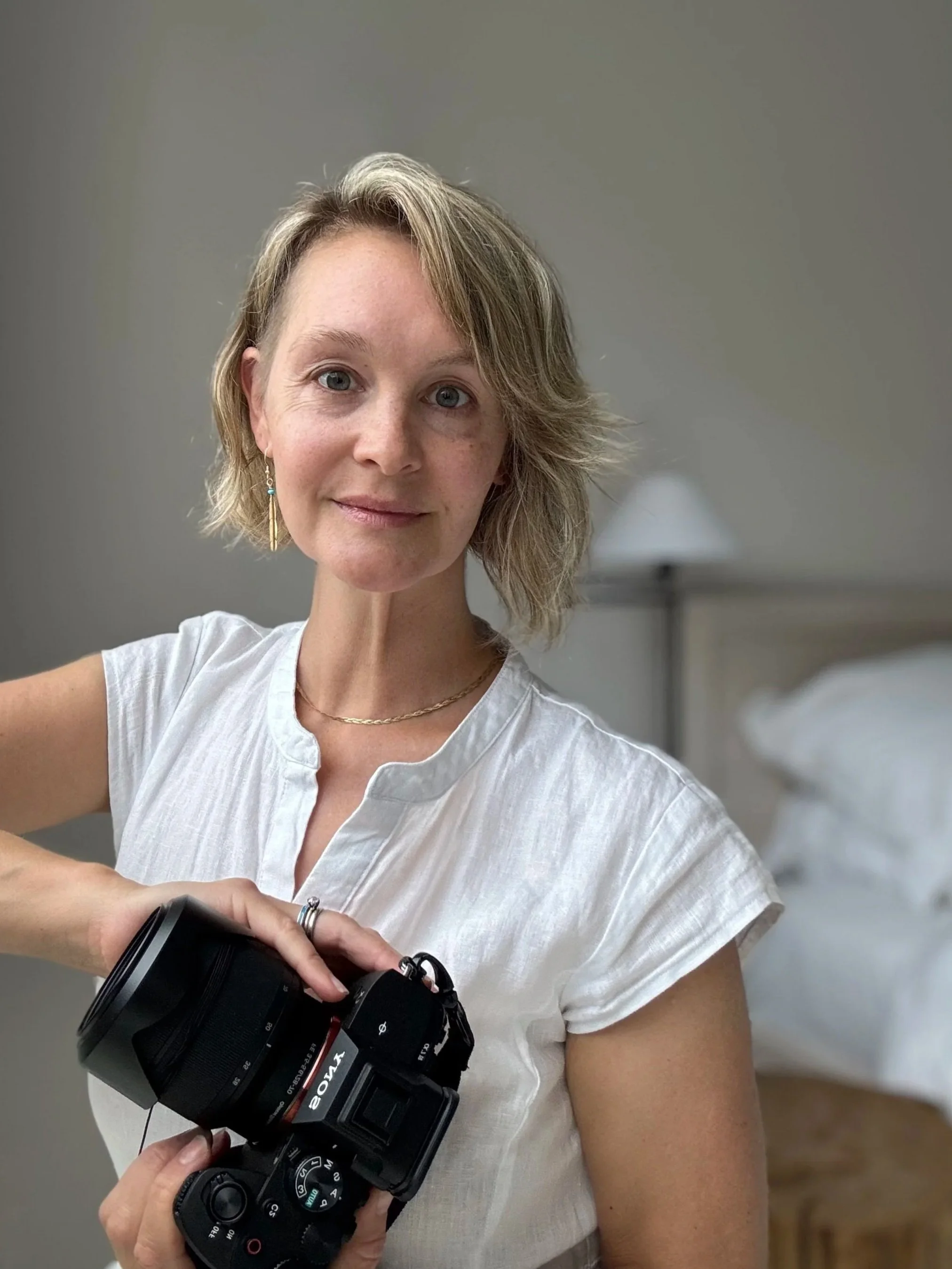 A woman with short, wavy blonde hair and blue eyes holding a camera in a room with a bed and a bedside lamp in the background.