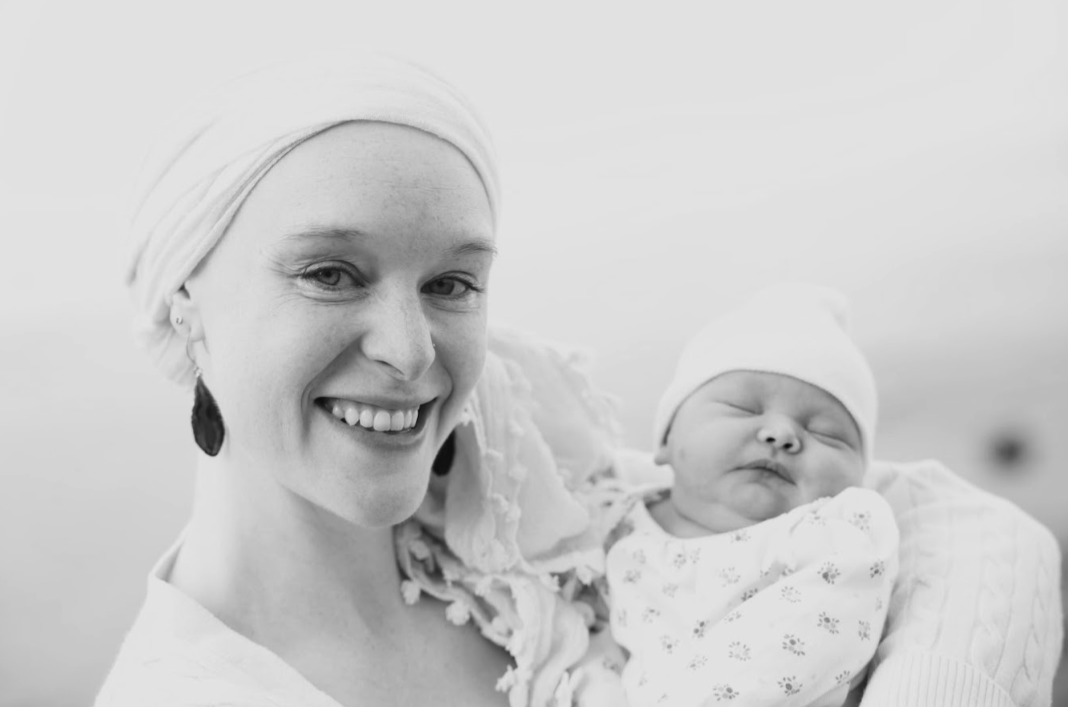 A woman with a head wrap and earrings smiling, holding a sleeping baby dressed in a patterned outfit and a hat.