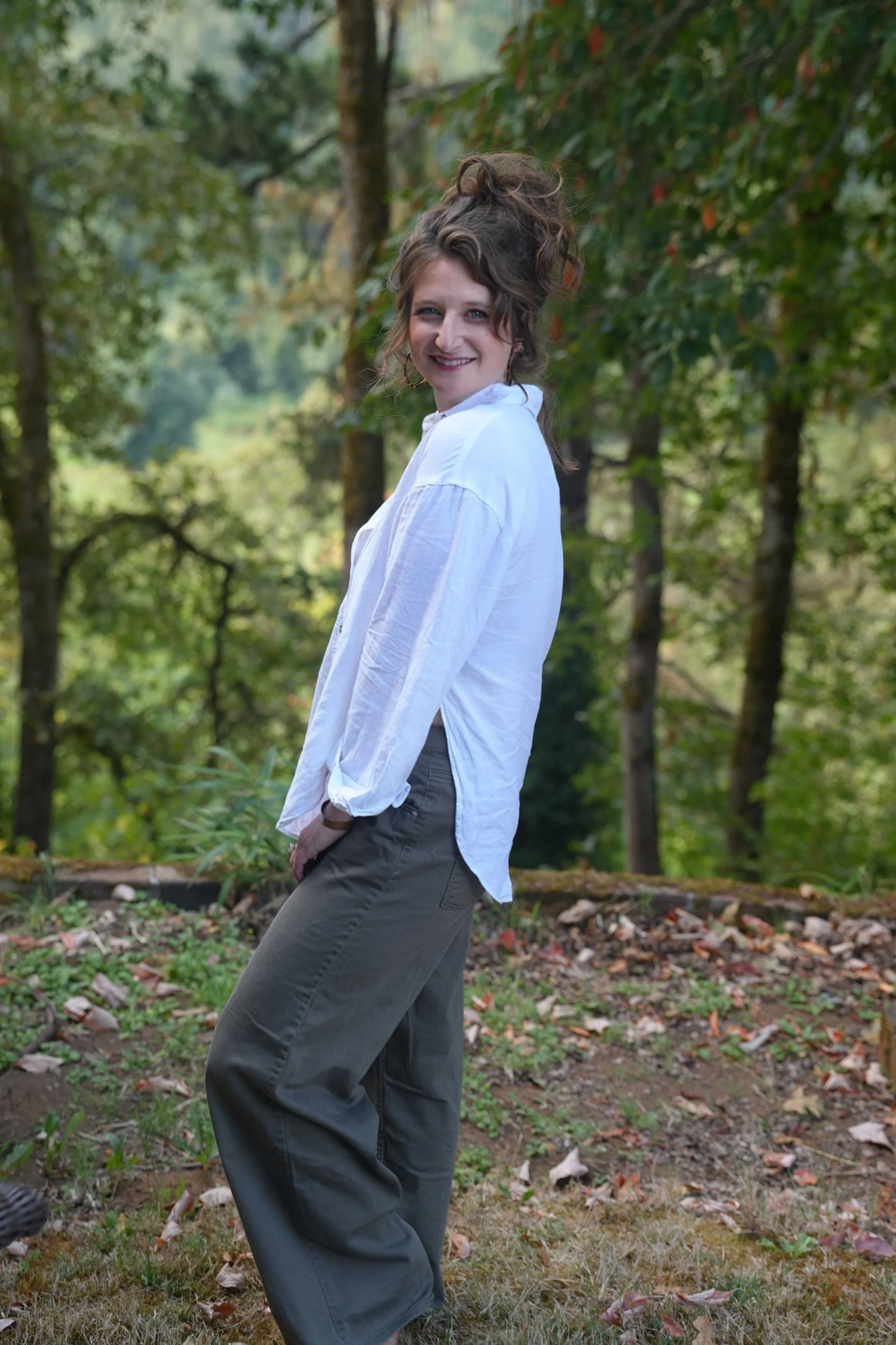 A woman with curly hair smiling outdoors in a forest, wearing a white shirt and gray pants.