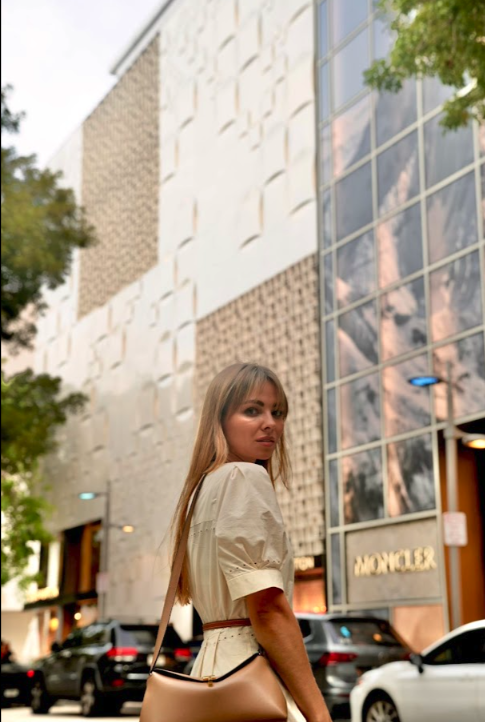 A woman with long blonde hair stands on a city street, turning to look back, with modern buildings and parked cars in the background.
