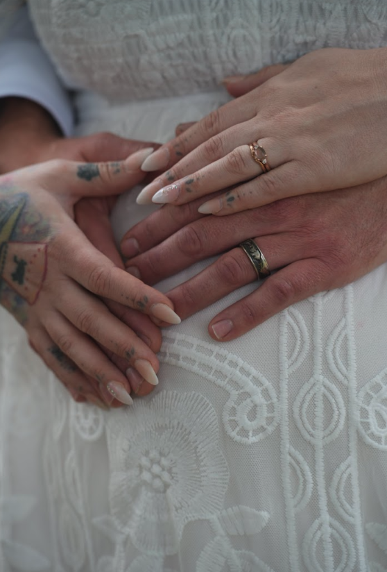 Two hands with rings and tattoos resting on a white textured fabric, symbolizing a close relationship or union.