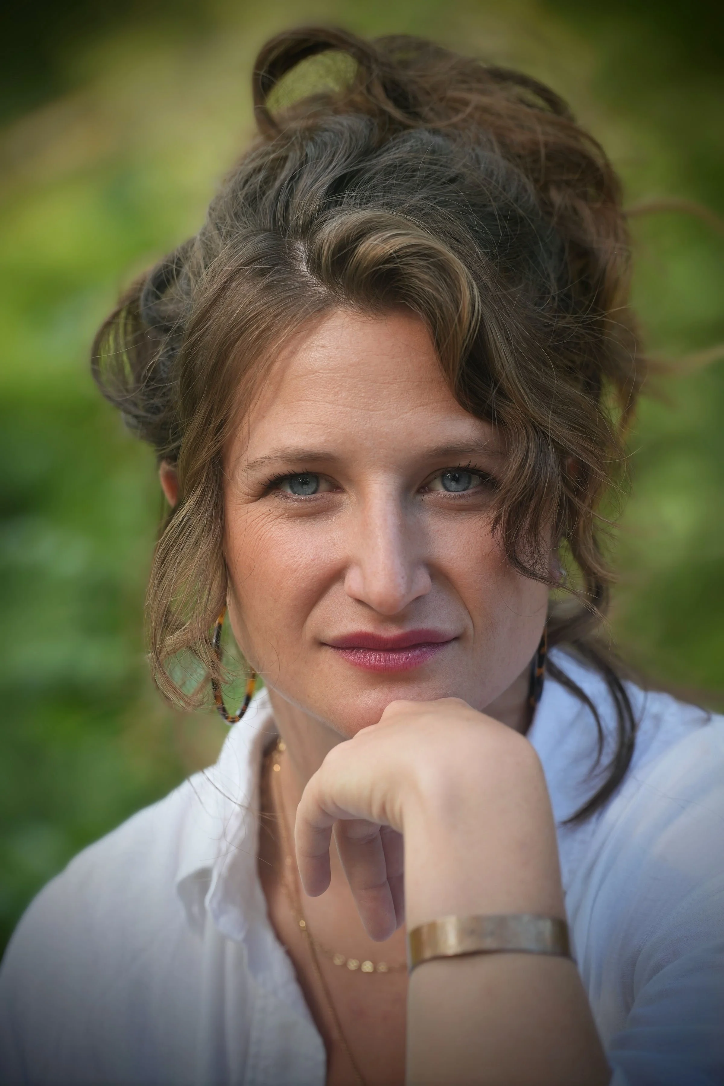 A woman with shoulder-length brown hair and blue eyes rests her chin on her hand, outdoors with a blurred green background.