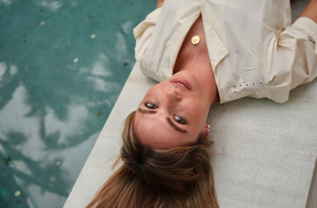 A woman with light brown hair and blue eyes lying on a white surface, wearing a beige jacket and a gold necklace, gazing upward.