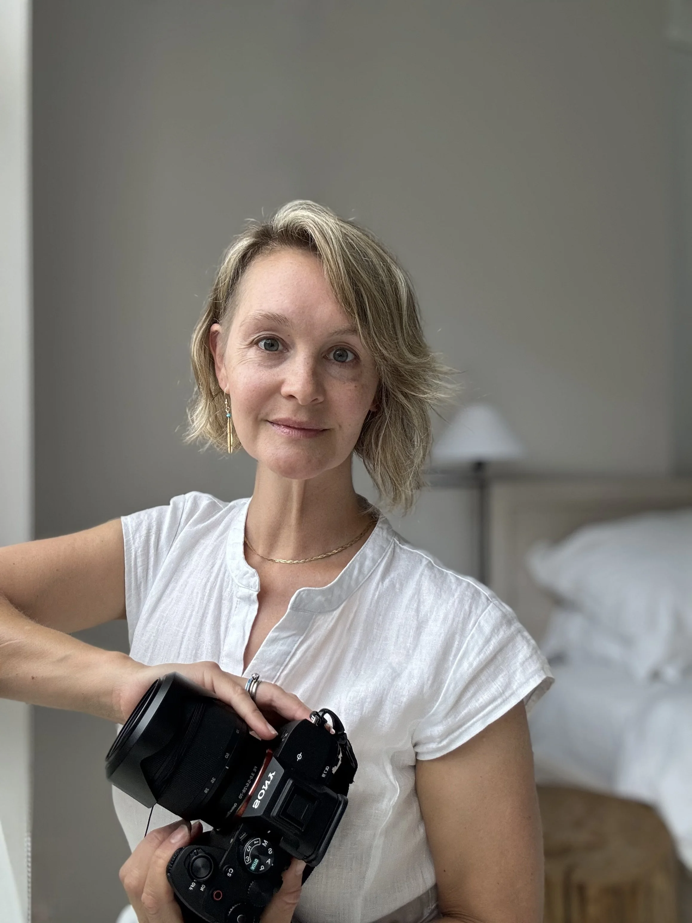 A woman with short blonde hair holding a Sony camera inside a room with a bed and a nightstand with a lamp in the background.