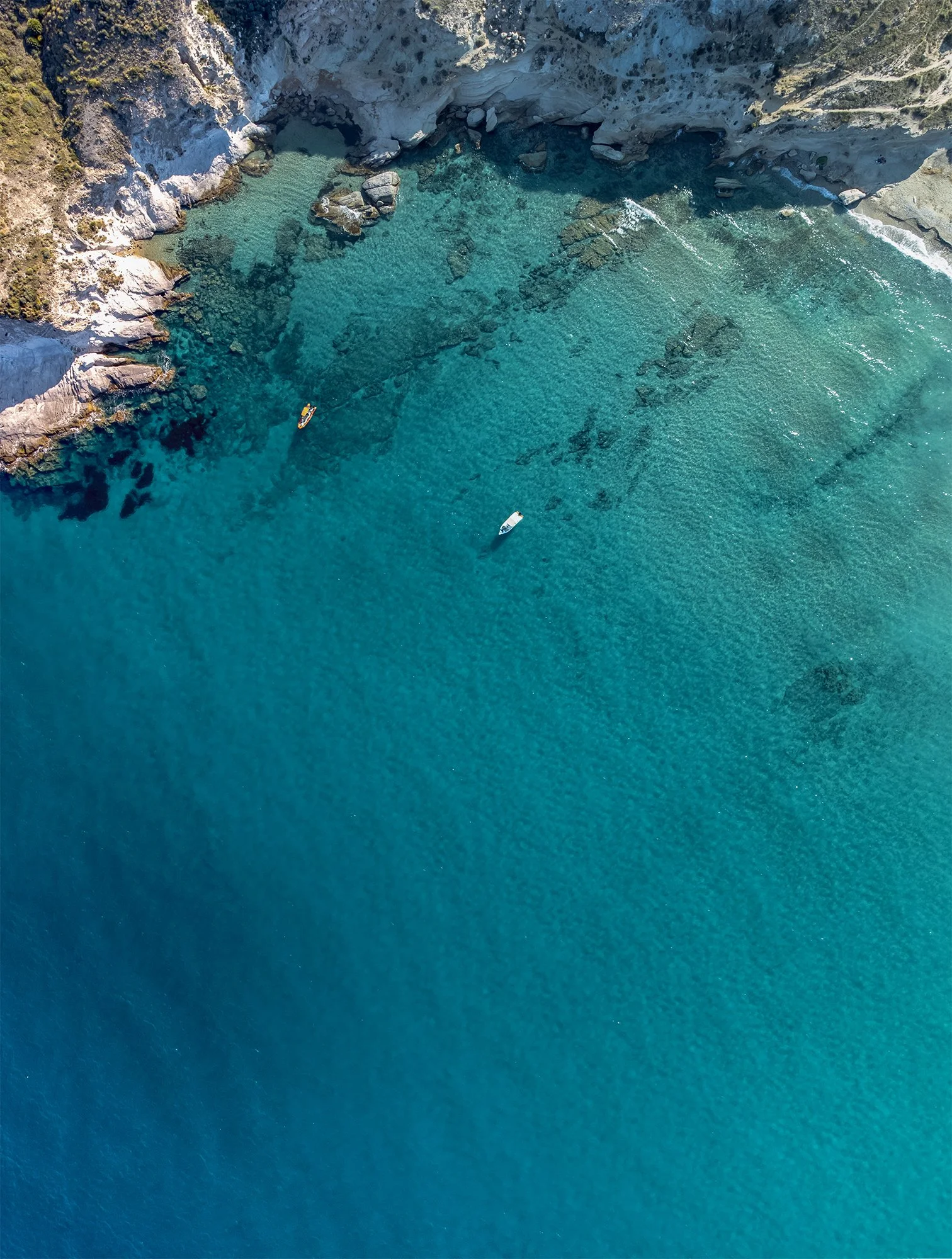 Aerial view of turquoise ocean water near rocky shoreline with two boats floating in the water.