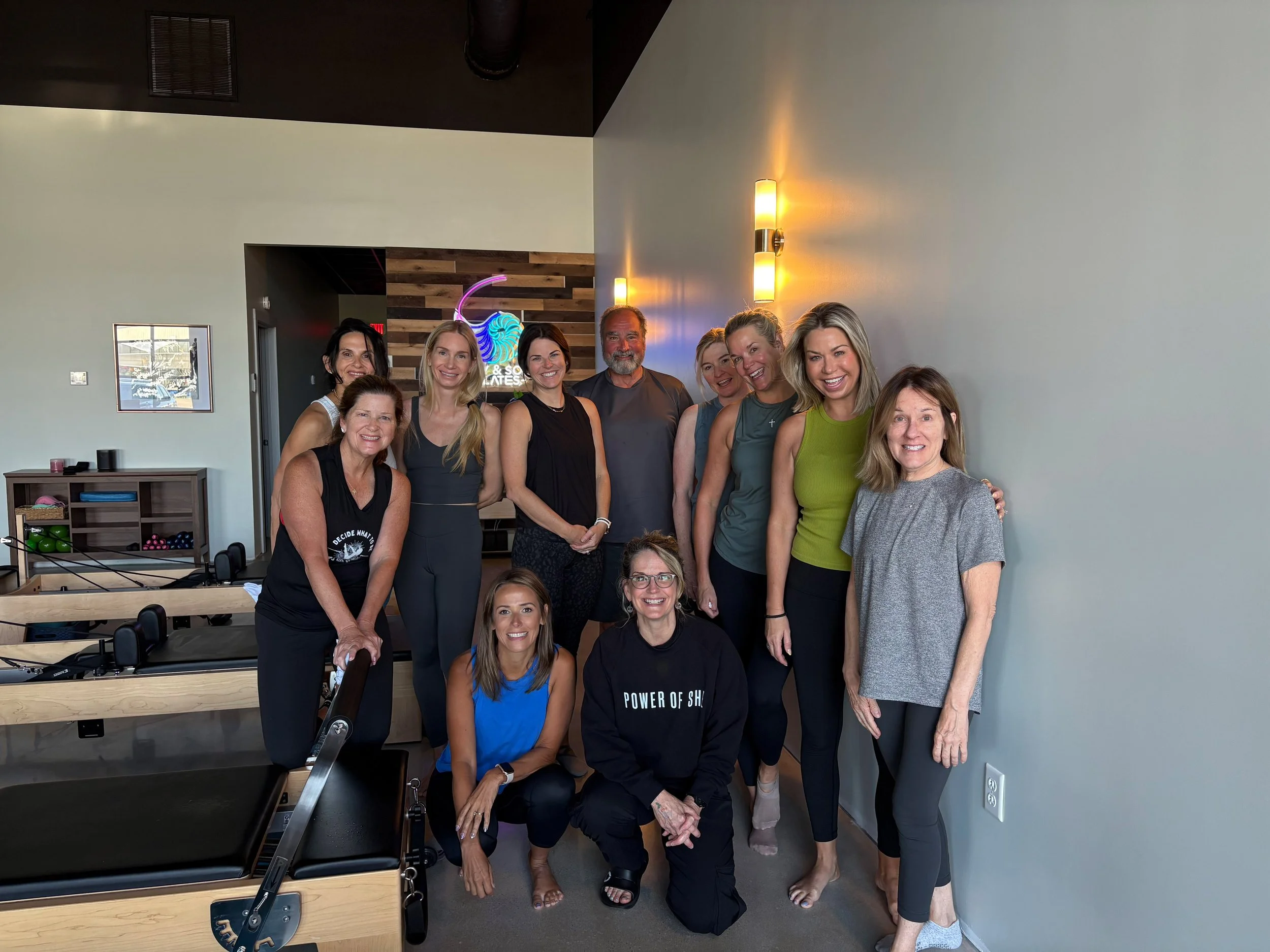 A group of twelve women and one man posing together at Body & Soul Pilates in Gluckstadt, MS, with low lighting and a neon sign in the background.