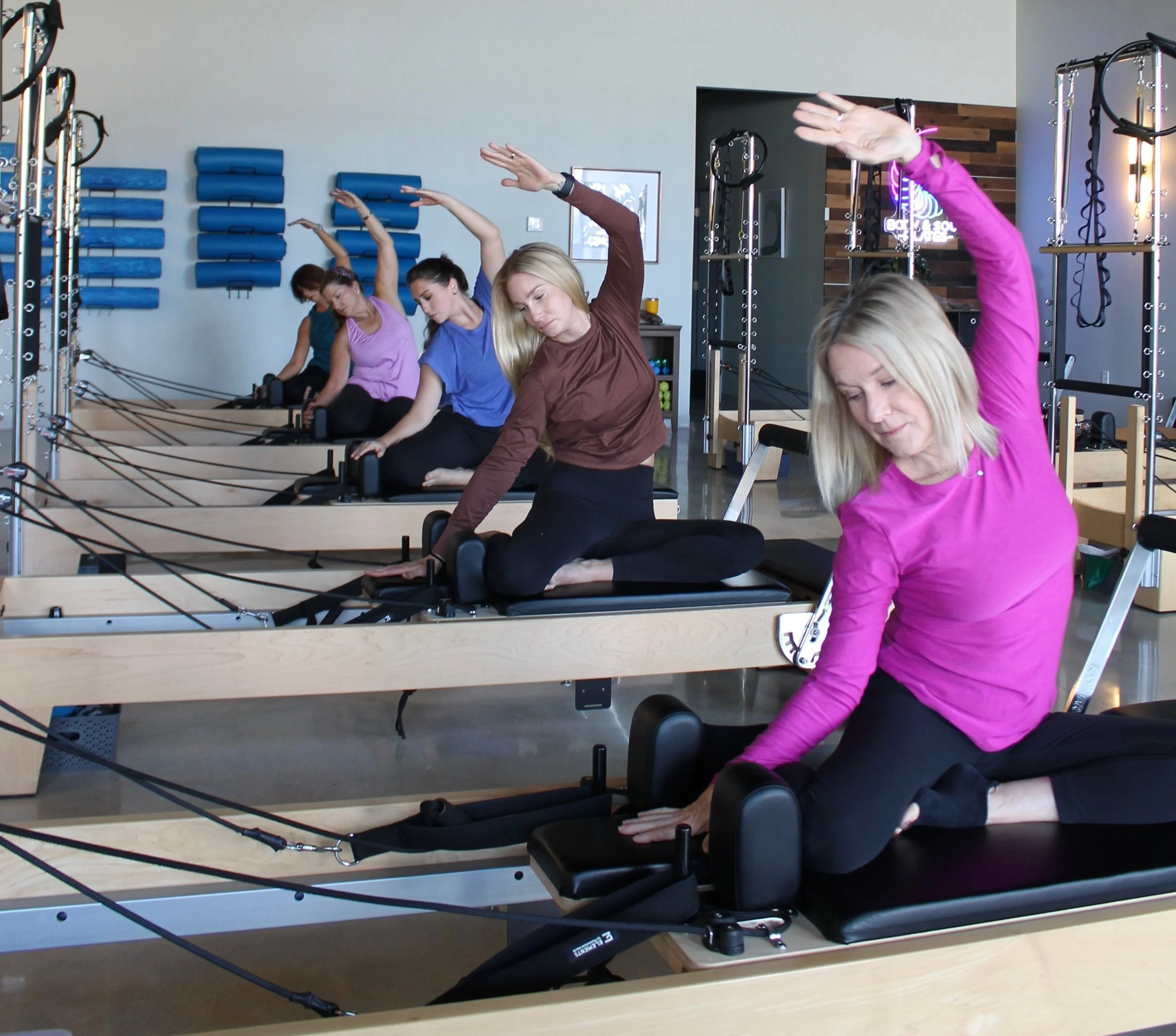 Group of women stretching on Pilates reformer machines in a fitness studio with blue foam rollers and gym equipment in the background.