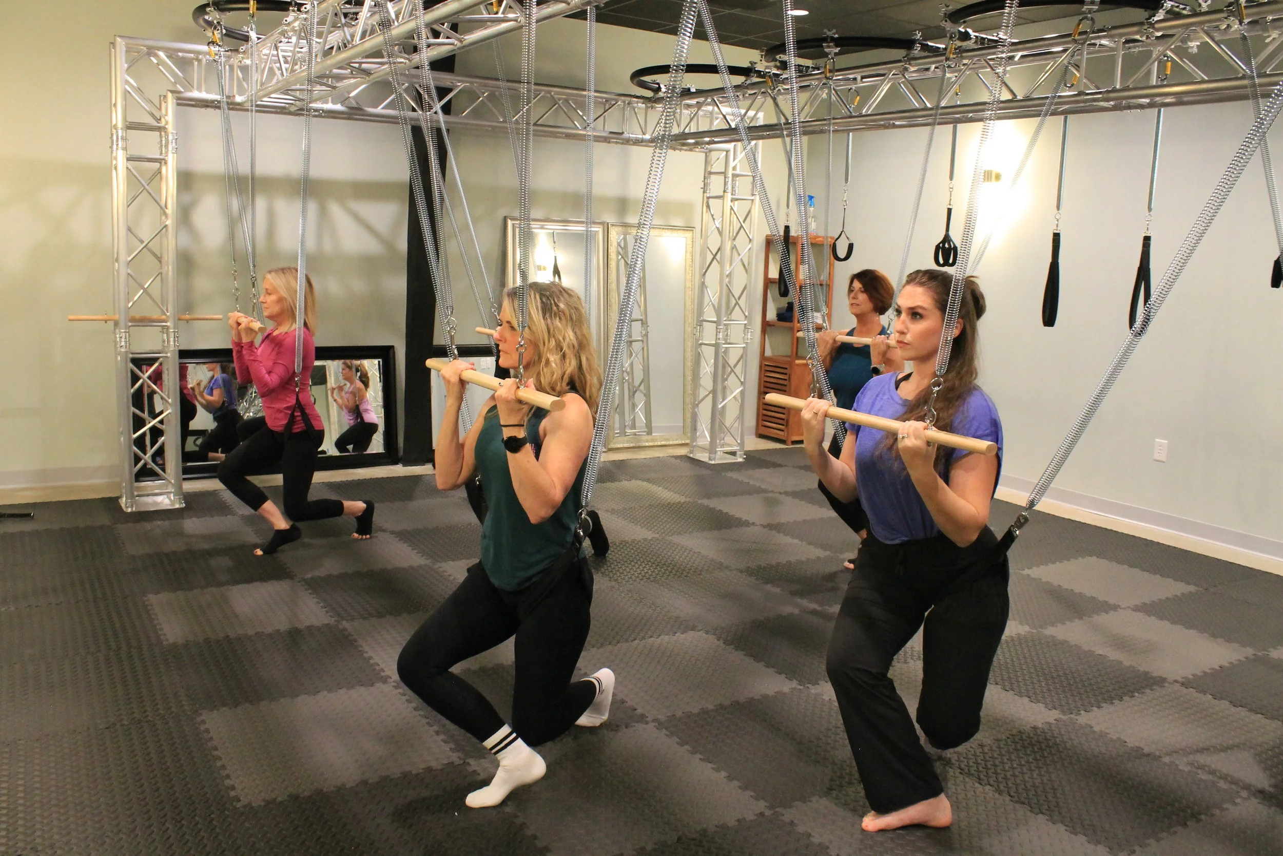 Four women are doing suspension training exercises with black straps attached to a metal frame in a fitness studio. They are in side lunge positions, holding onto the straps for support.