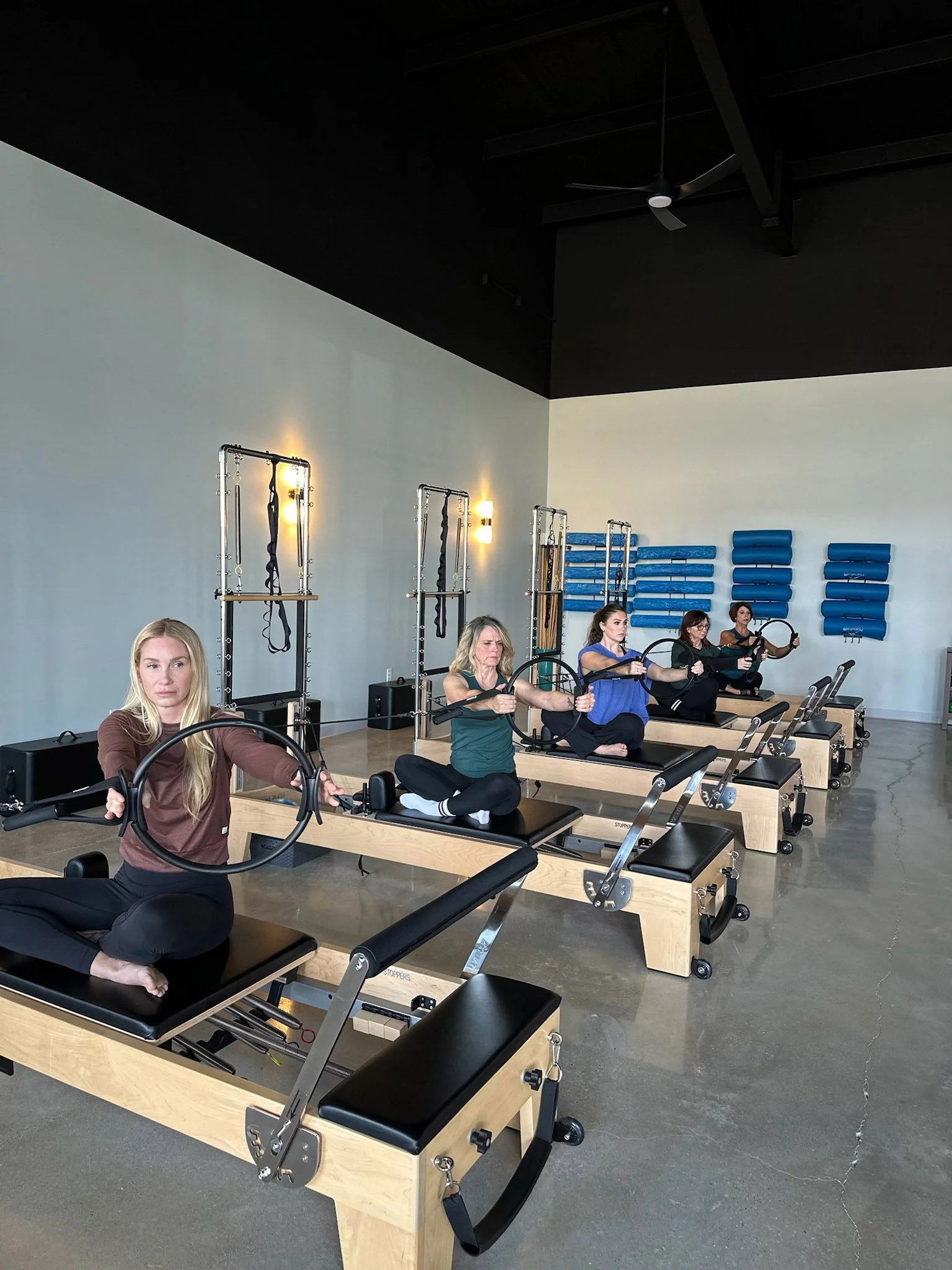 A group of women practicing Pilates on reformer machines at Body & Soul Pilates in Gluckstadt, MS with blue wall-mounted equipment and a black ceiling fan.