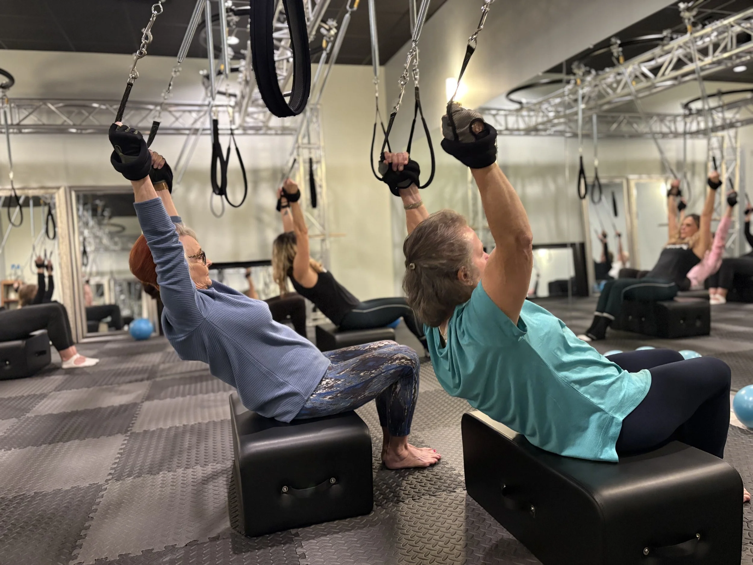 Older women and men participating in a fitness class at a gym, performing seated yoga or stretching exercises with their arms raised and holding suspension straps while seated on black benches.