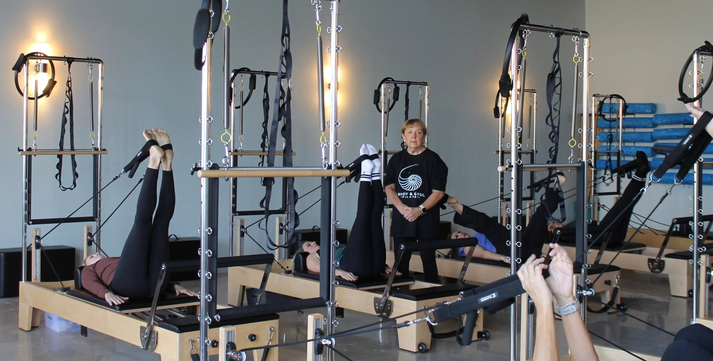Body & Soul Pilates studio with people exercising on reformer machines, instructor supervising, light-colored walls, stack of blue mats in background.