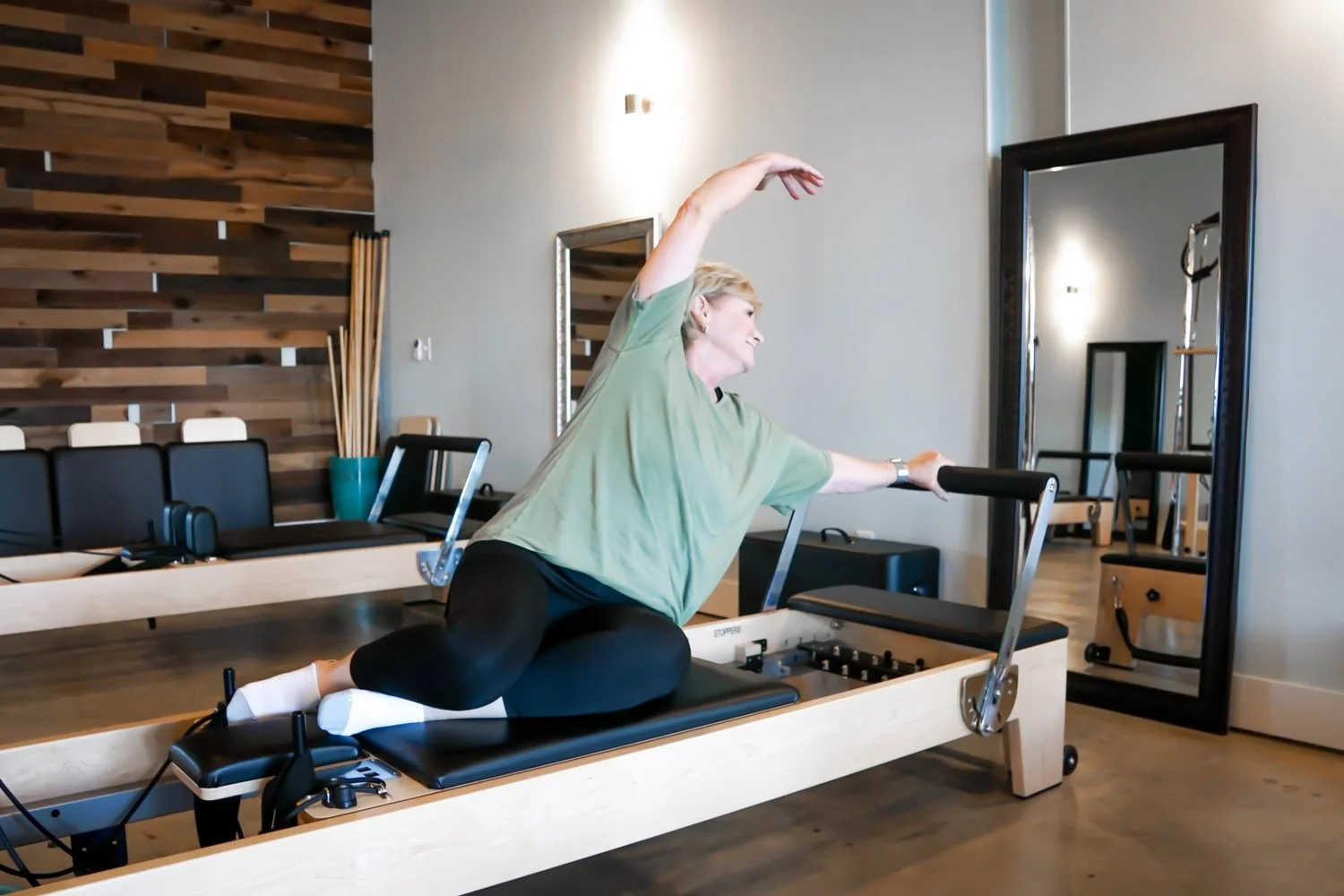 Woman exercising on a reformer machine at Body & Soul Pilates in Gluckstadt, MS, performing a side stretch with one arm raised overhead and the other on the reformer