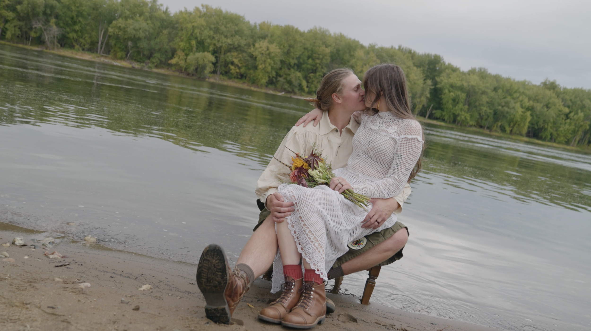 A couple sitting on a small stool near a river, sharing a kiss, with the woman holding a bouquet of flowers, and the man wearing hiking boots, a beige shirt, and shorts. The woman is in a white lace dress with long sleeves.