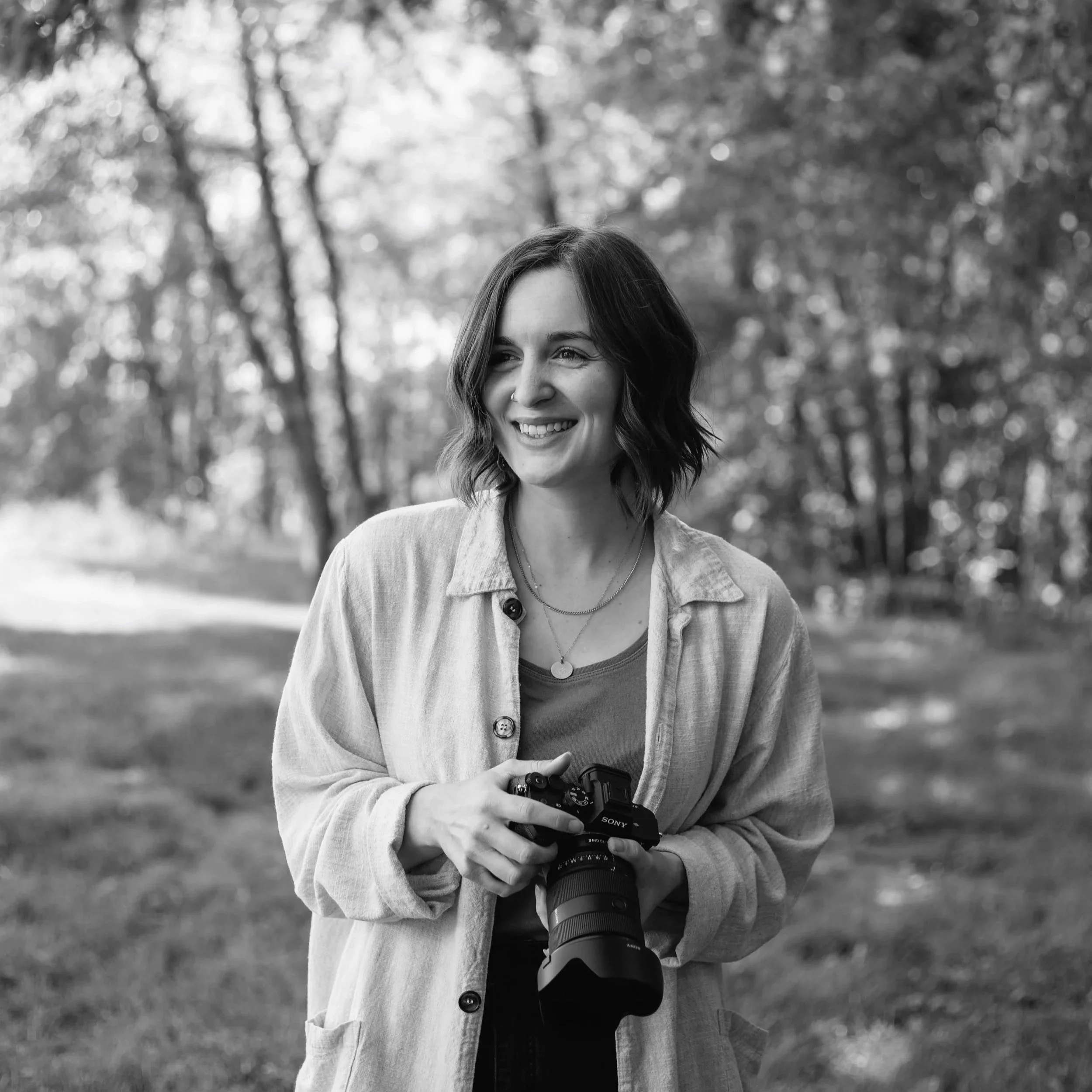 A woman with shoulder-length wavy hair smiling and holding a camera outdoors in a park with trees in the background.