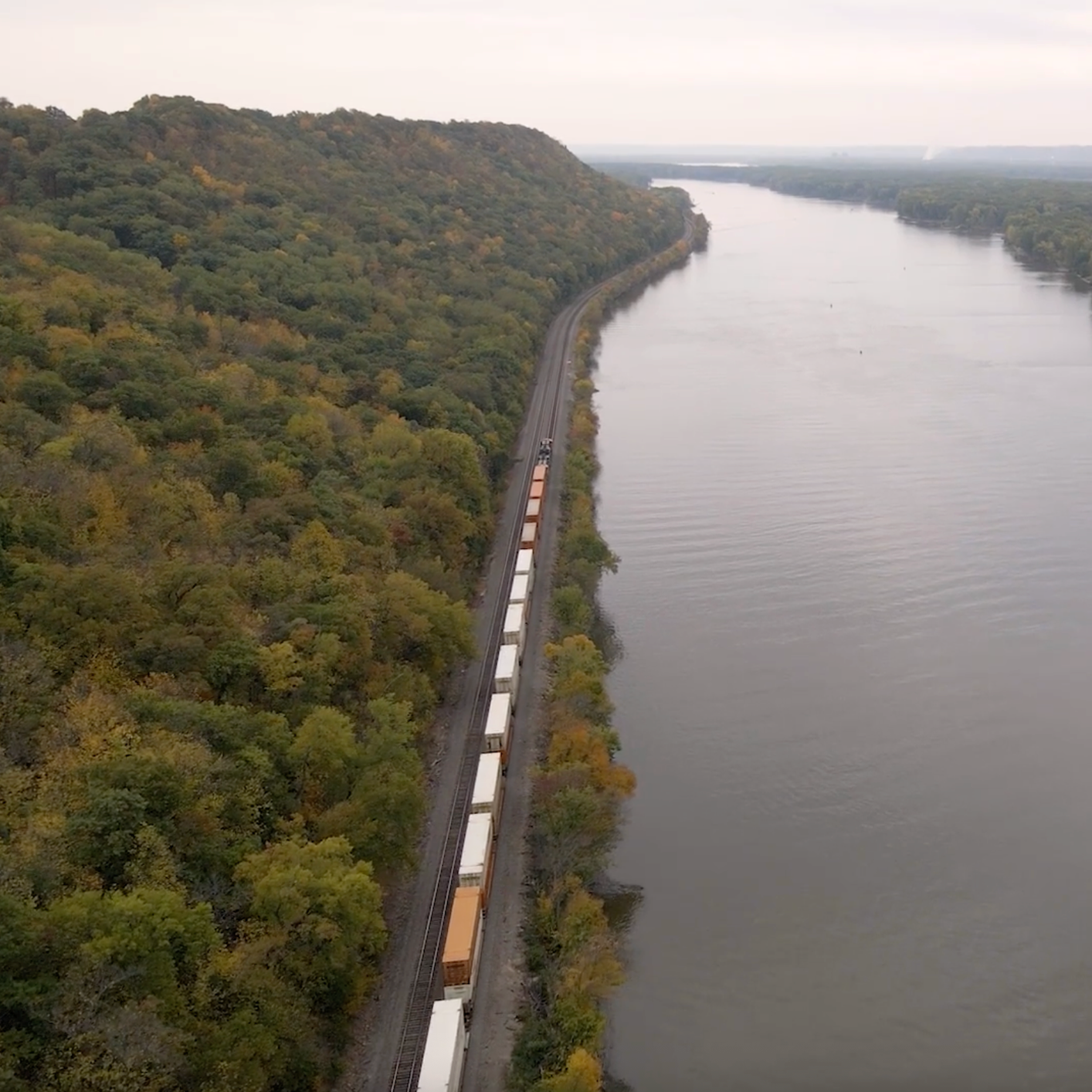 An aerial view of a train traveling along a railway track beside a wide river, with a forested hill on the left and open water on the right.
