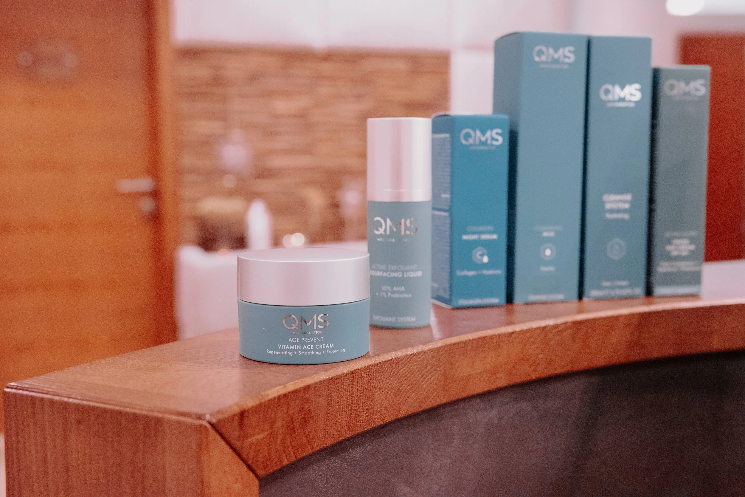 Mehrere QMS-Hautpflegeprodukte, darunter eine Creme und ein Serum, auf einem Holztheke in einem Raum mit warmem Licht.
