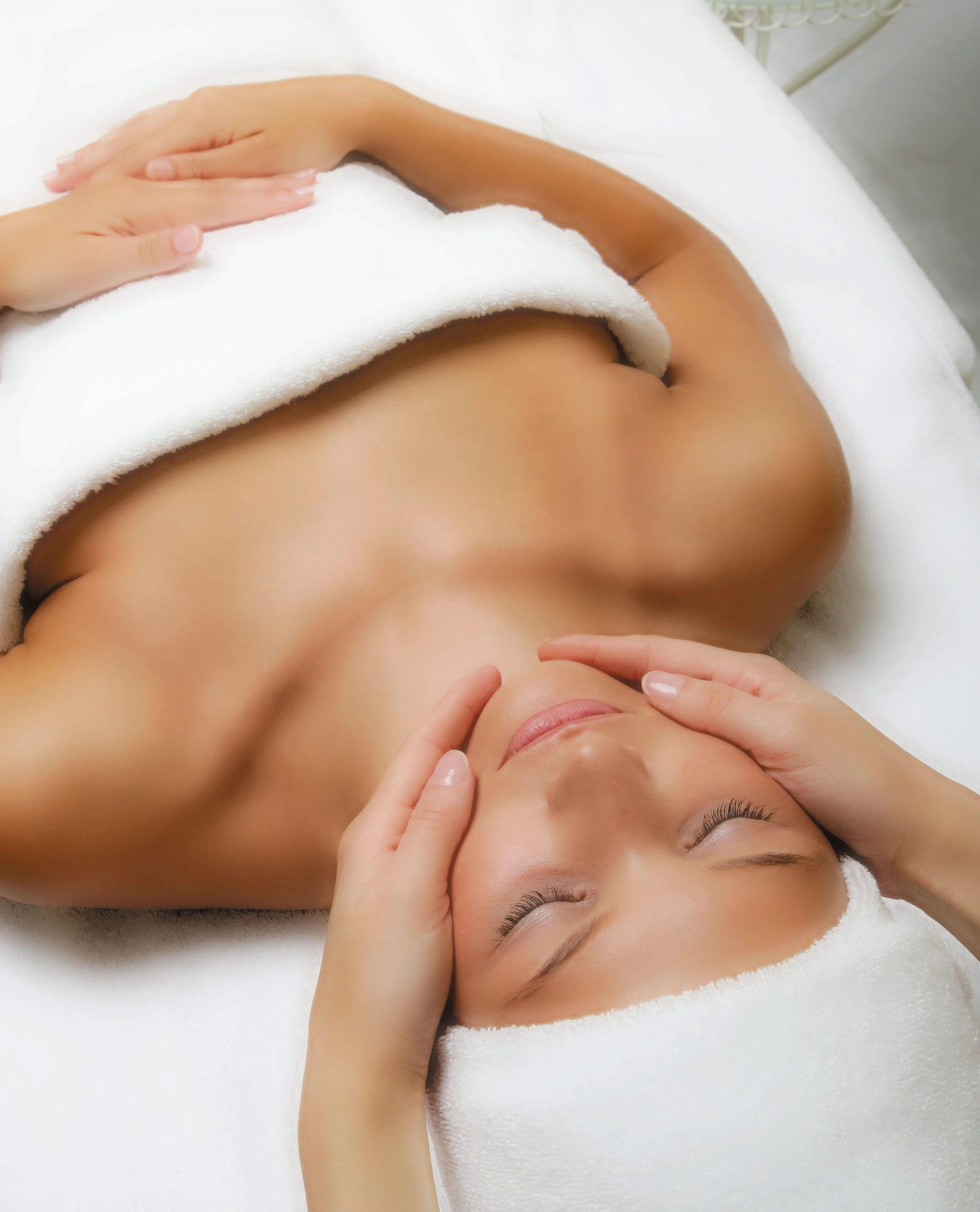 Frau erhält eine Gesichtsmassage, liegend auf einer Massagebank, mit weißen Handtüchern bedeckt.