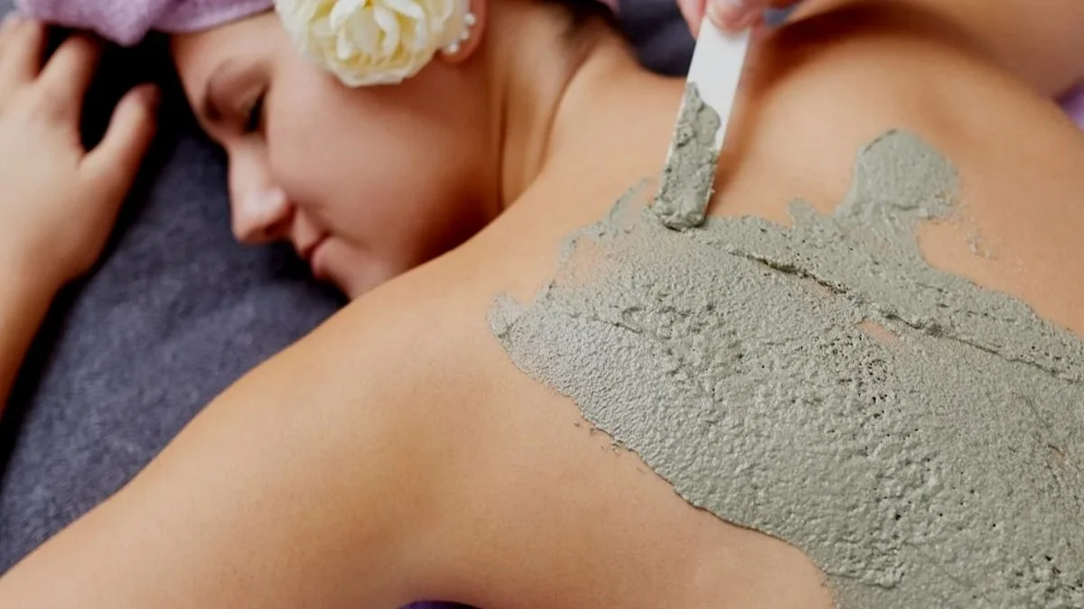 Frau liegt entspannt mit Blumen im Haar, während sie mit einem Spachtel grauen Mörtel auf ihre Schulter aufträgt.