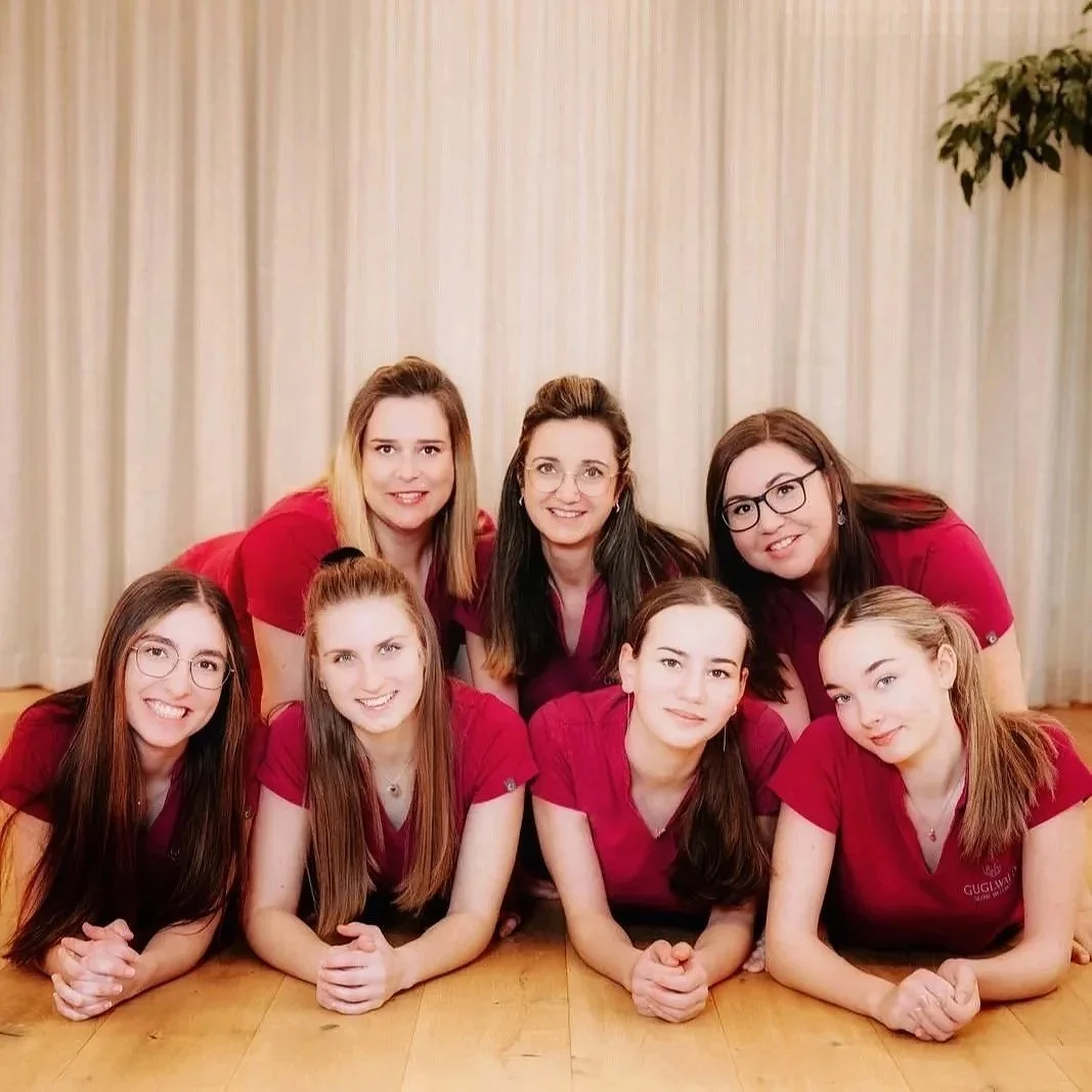Sieben Frauen in roten T-Shirts sitzen und knien auf einem Holzboden vor einem weißen Vorhang, lächelnd für ein Gruppenfoto.