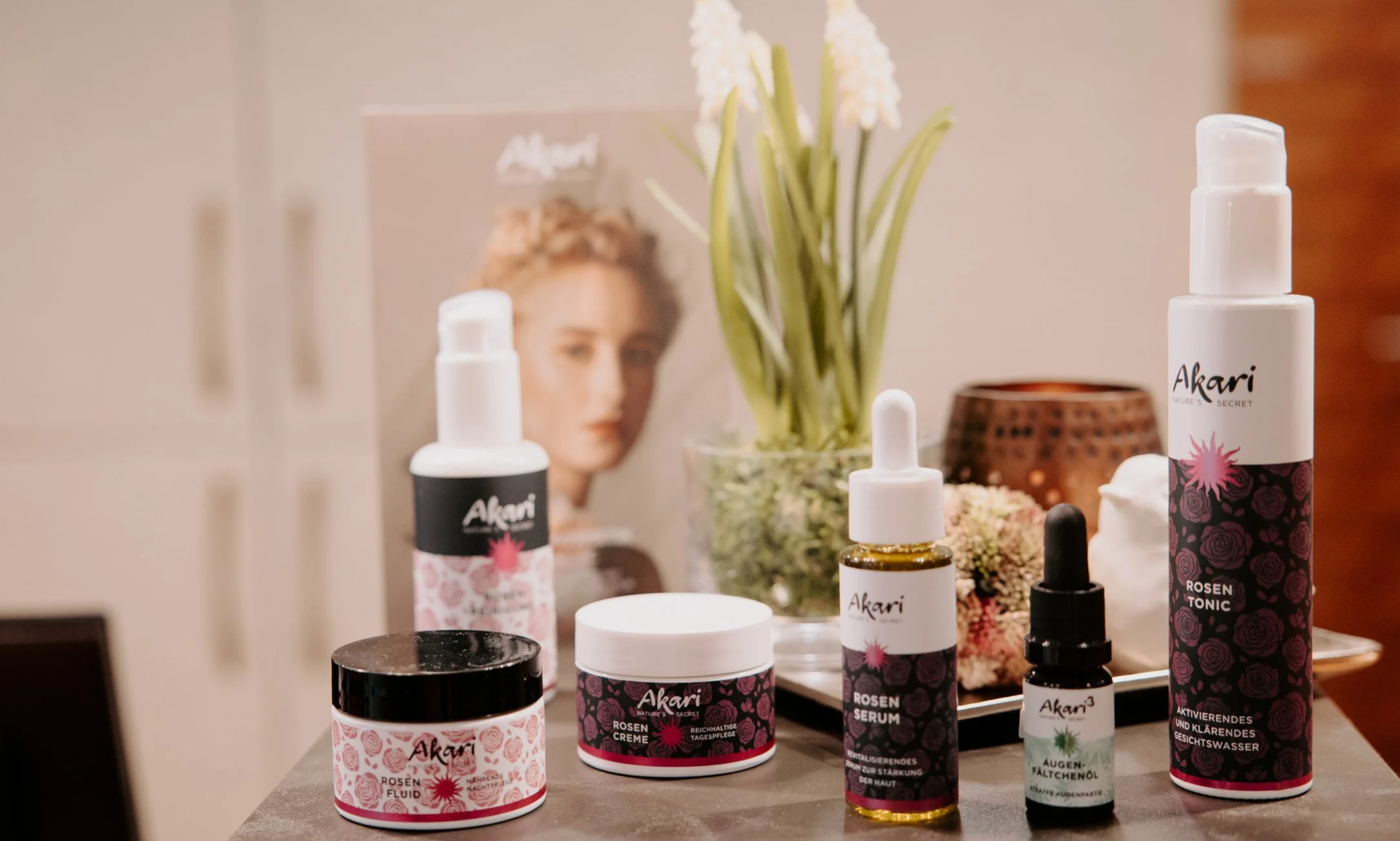 Hautpflegeprodukte mit Rosen, eine Vase mit weißen Blumen, ein Magazin, ein Kerzenhalter, und eine dunkle Oberfläche im Hintergrund.