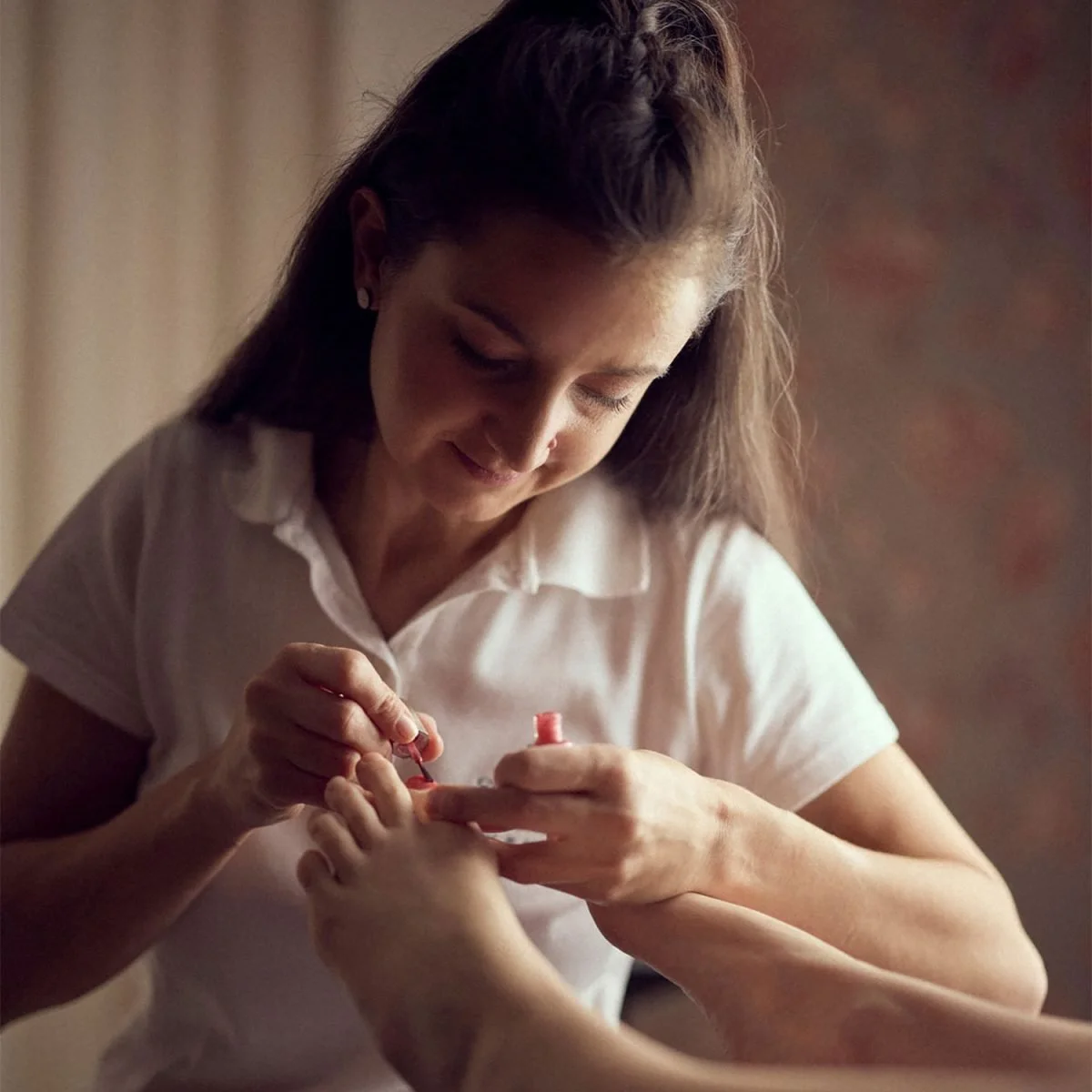 Frau, die Fußnägel lackiert, trägt weiße Bluse, sitzt in gut beleuchtetem Raum.