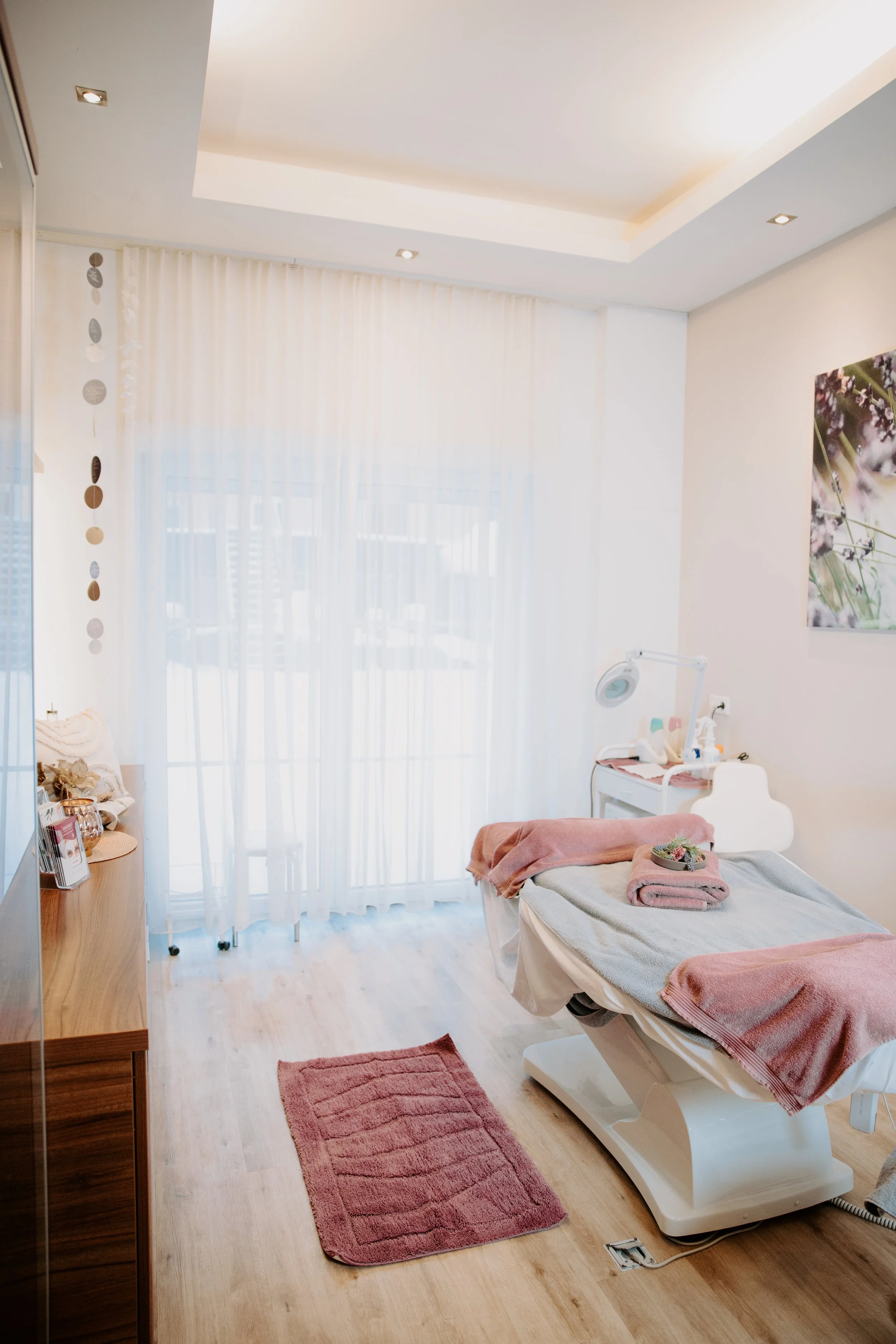 Ein Behandlungsraum mit Liege, Tisch, Medizin- und Pflegeutensilien, Vorhängen, Holzboden und modernen Lichtquellen.