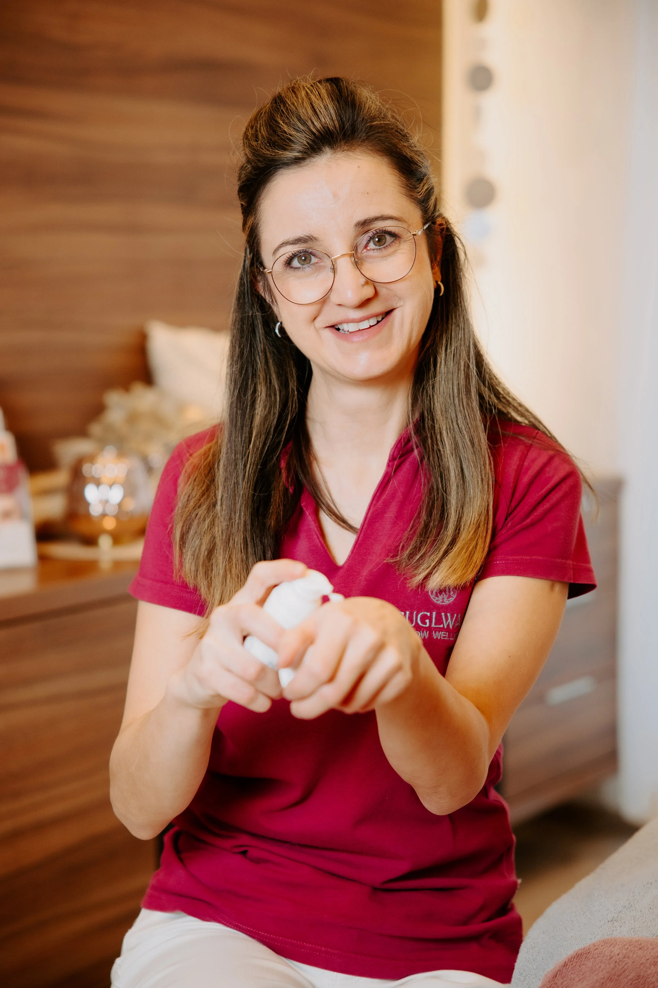 Frau mit Brille, die ein Hautpflegeprodukt auf ihre Hand aufträgt, lächelnd in einem gemütlichen Raum.