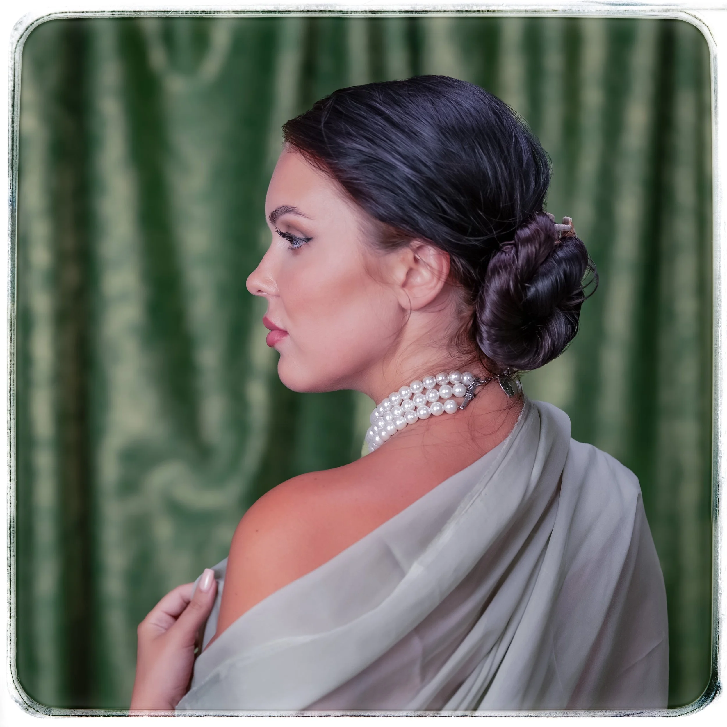 Eine Frau mit dunklen, glatten Haaren, die zu einem Knoten zusammengebunden sind, trägt mehrere Perlenketten und einen leichten, grauen Schal. Sie steht vor einem grünen, verschwommenen Hintergrund und schaut nach links.