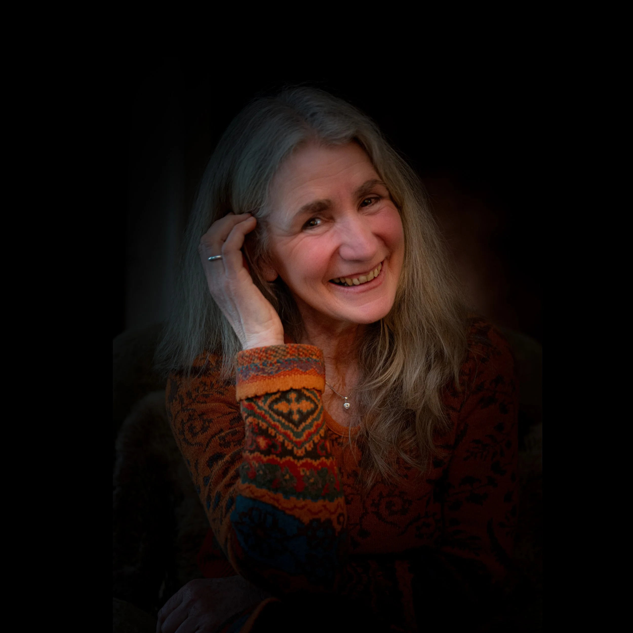Ein lächelndes älteres Frauenporträt mit grauen Haaren, die lächelnd den Kopf leicht neigt und eine Hand ans Ohr legt, getragen mit einem bunten, gemusterten Ärmel. Dunkler, unscharfer Hintergrund.