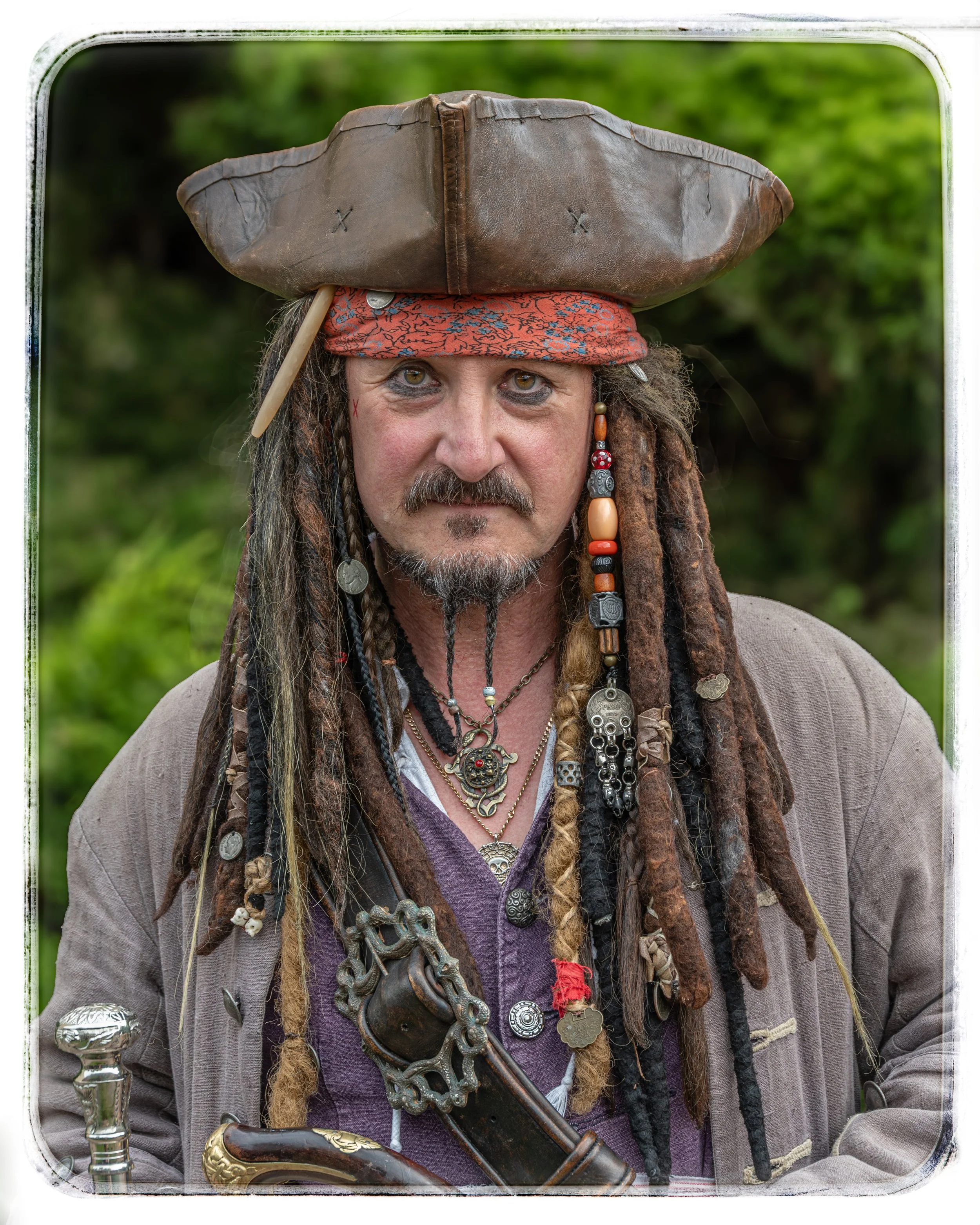 Ein Mann mit Dreadlocks, Piratenkostüm, trug einen braunen Piratenhut, eine rote Bandana, und viele Halsketten, Accessoires und einen Degen, in einer grünen, bewaldeten Umgebung.