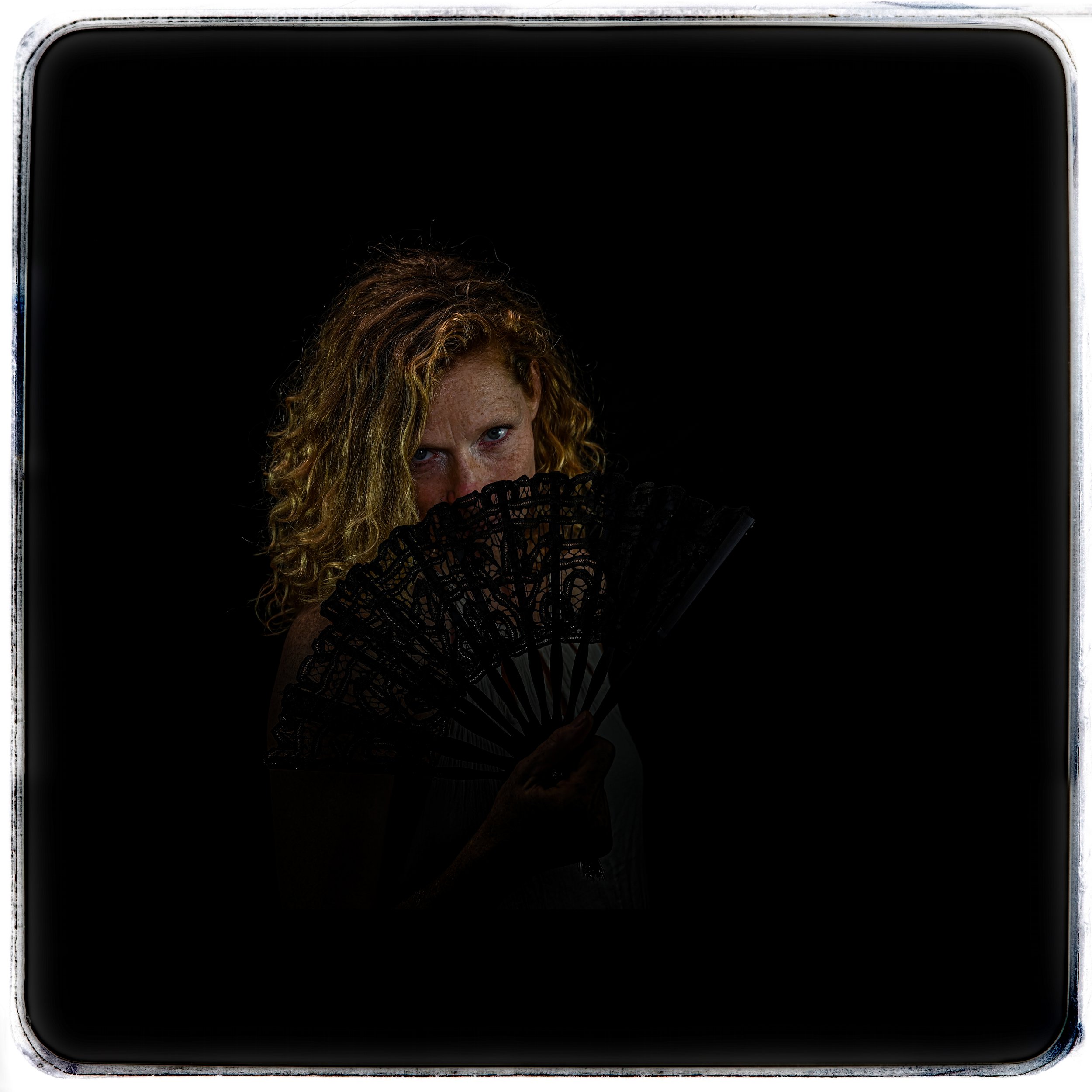 Frau mit roten, lockigen Haaren hält ein schwarzes Fächer vor das Gesicht, nur die Augen sichtbar, vor schwarzem Hintergrund.