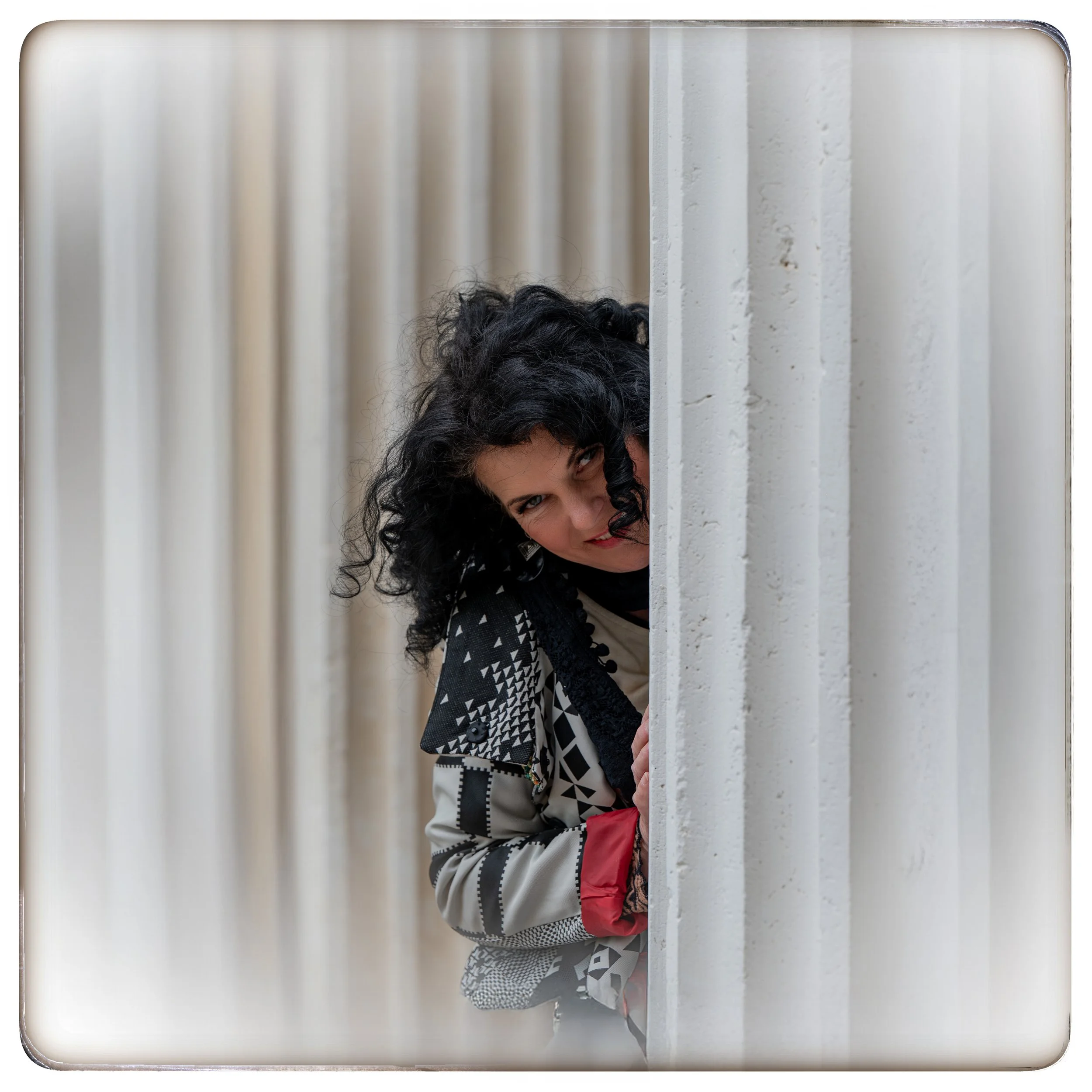 Frau mit schwarzen, lockigen Haaren, die hinter einer weißen Wand hervorschaut und lächelt.