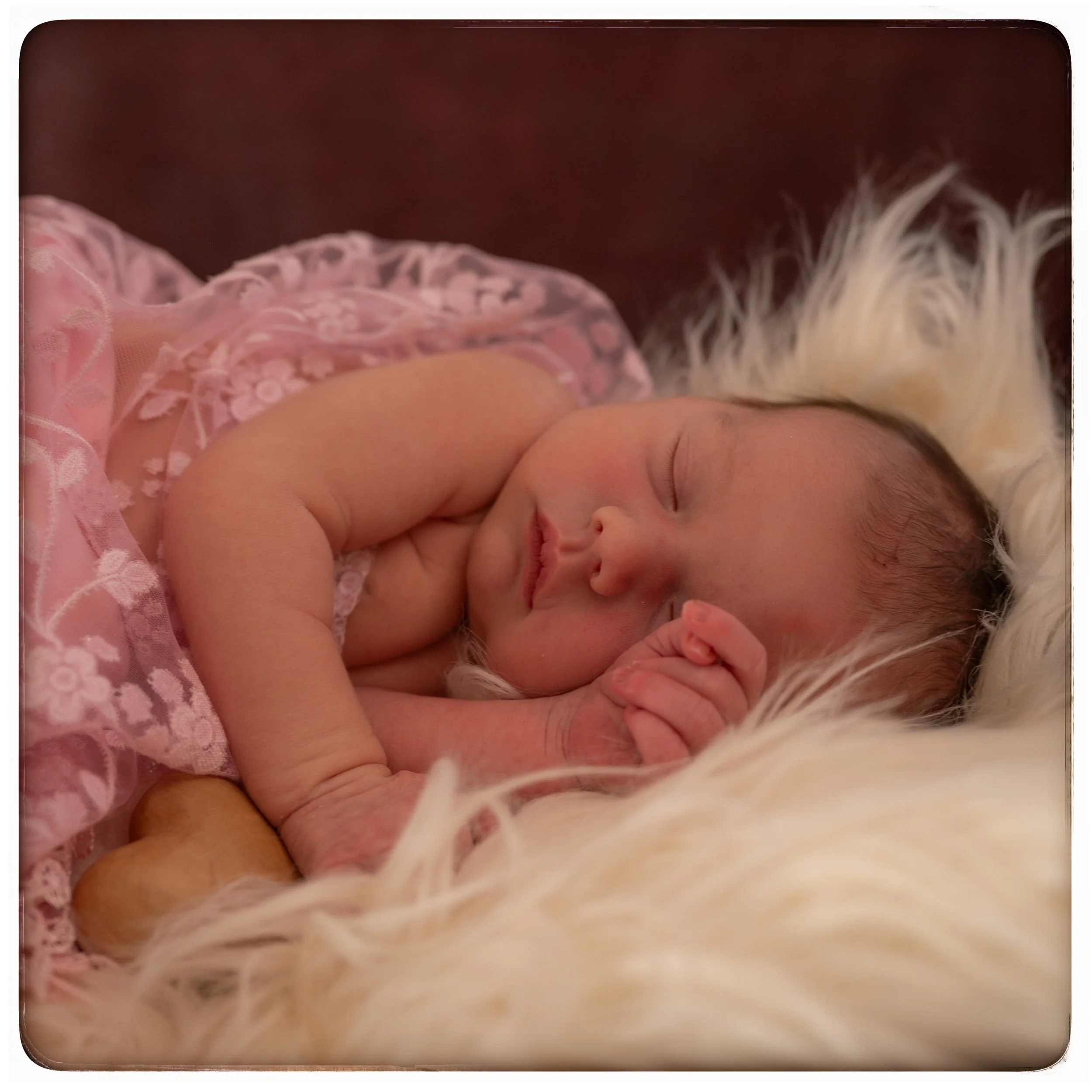 Ein neugeborenes Mädchen schläft auf einem weißen, pelzigen Kissen, trägt ein rosa, Spitzen- Baby-Kleid, mit geschlossenen Augen und einer Hand in der Nähe ihres Gesichts.