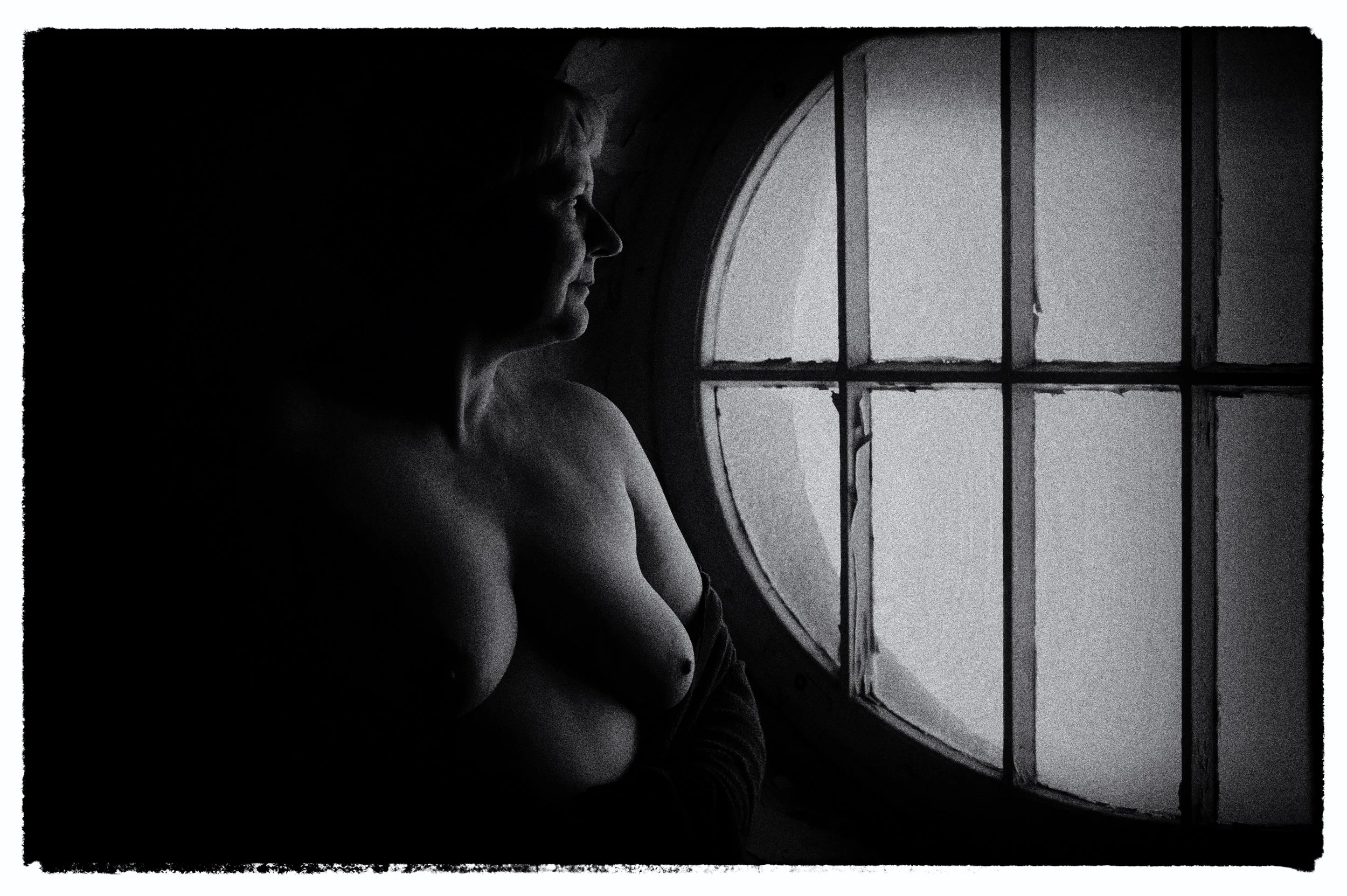 Eine nackte Frau sitzt am Fenster und schaut nach draußen in einem Raum mit dunklem Ambiente, schwarz-weißes Bild.